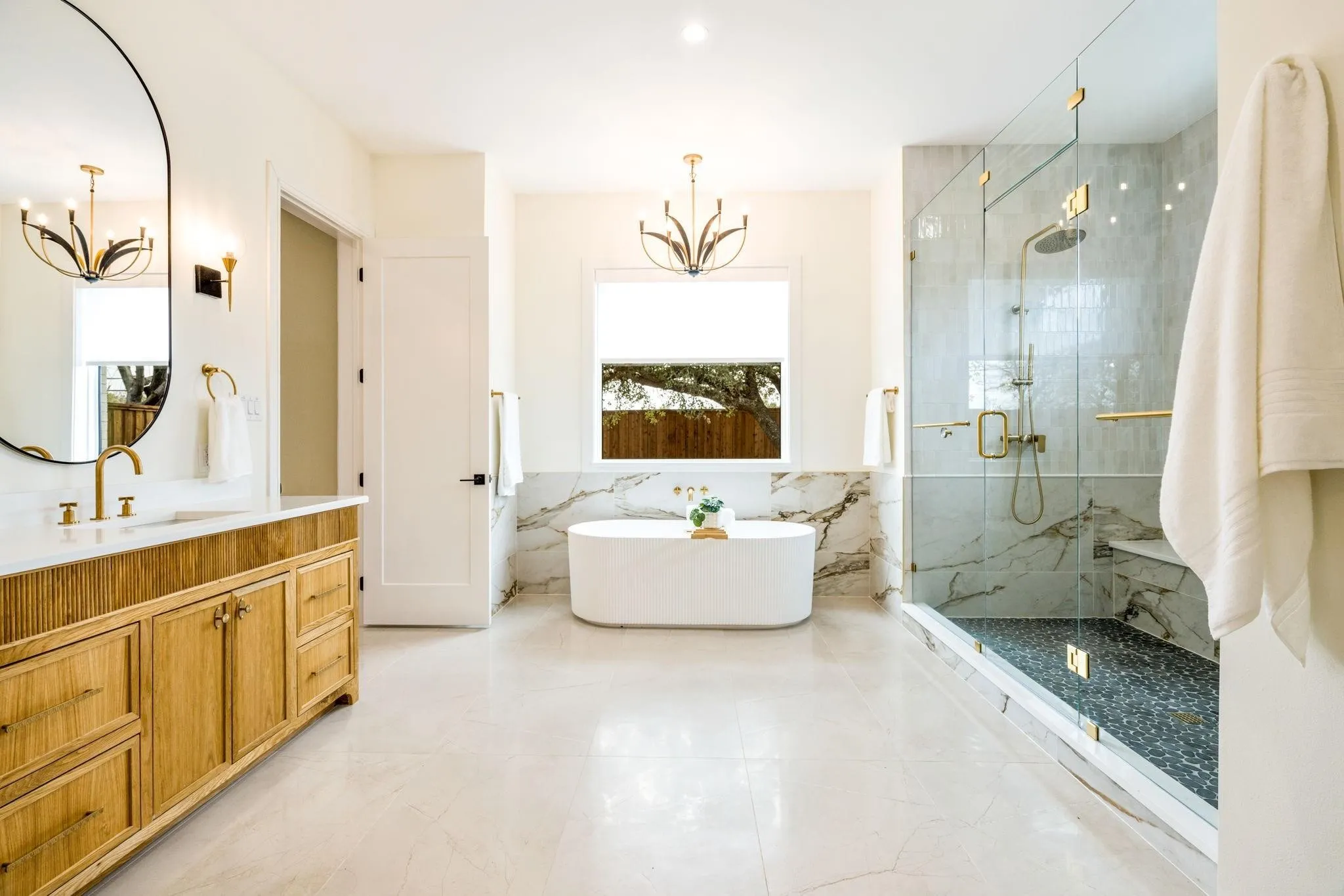Freestanding oval bathtub, glass-enclosed shower with a built-in bench, wood-finish vanity with a stone countertop, and an oval wall mirror