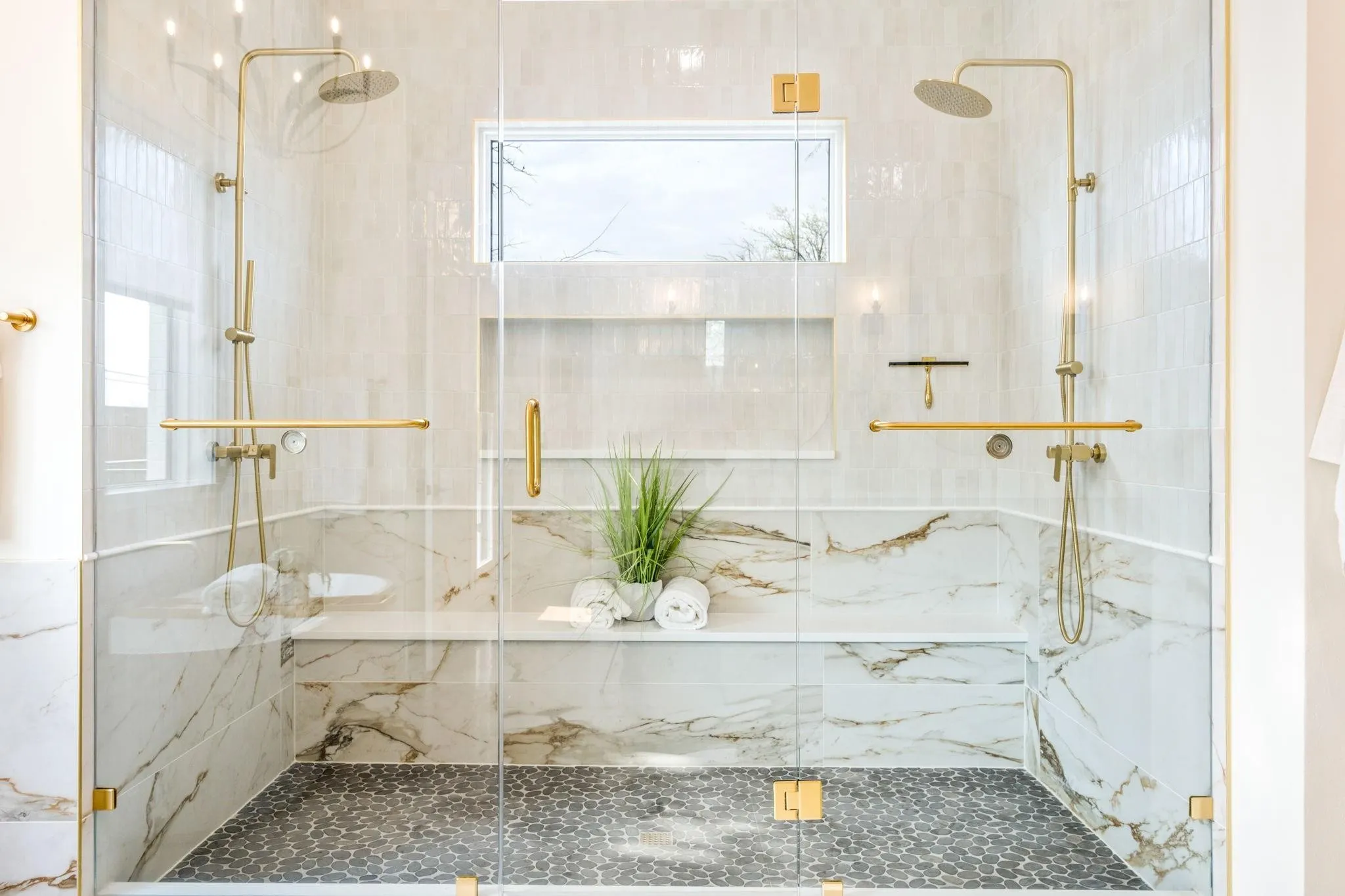 Dual rain shower heads with handheld sprayers, glass enclosure, built-in bench, marble-finish wall tiles, and pebble-tile flooring