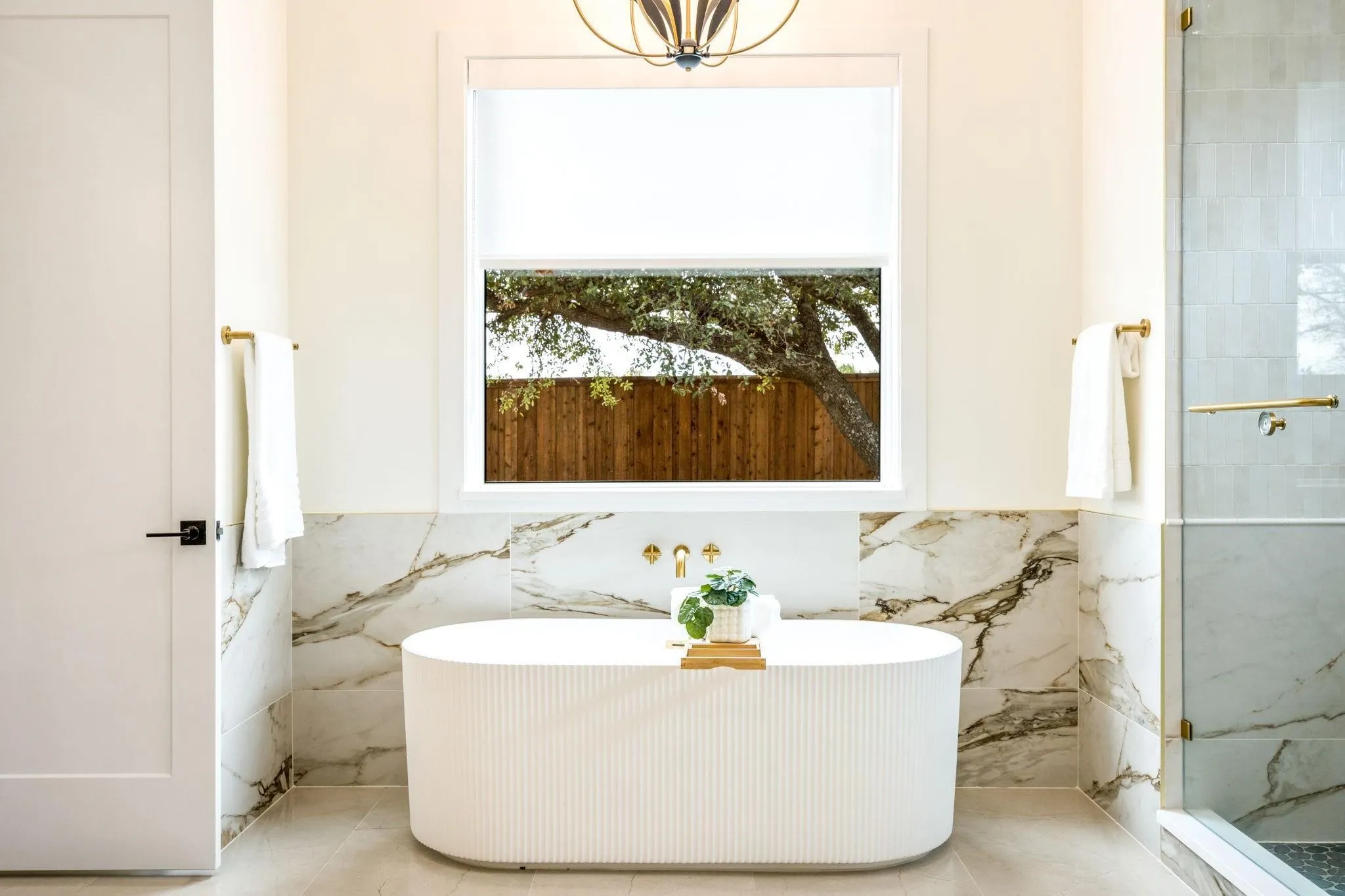 Freestanding soaking tub with a wall-mounted faucet, complemented by large format tile with natural veining, and a glass-enclosed shower with brass hardware