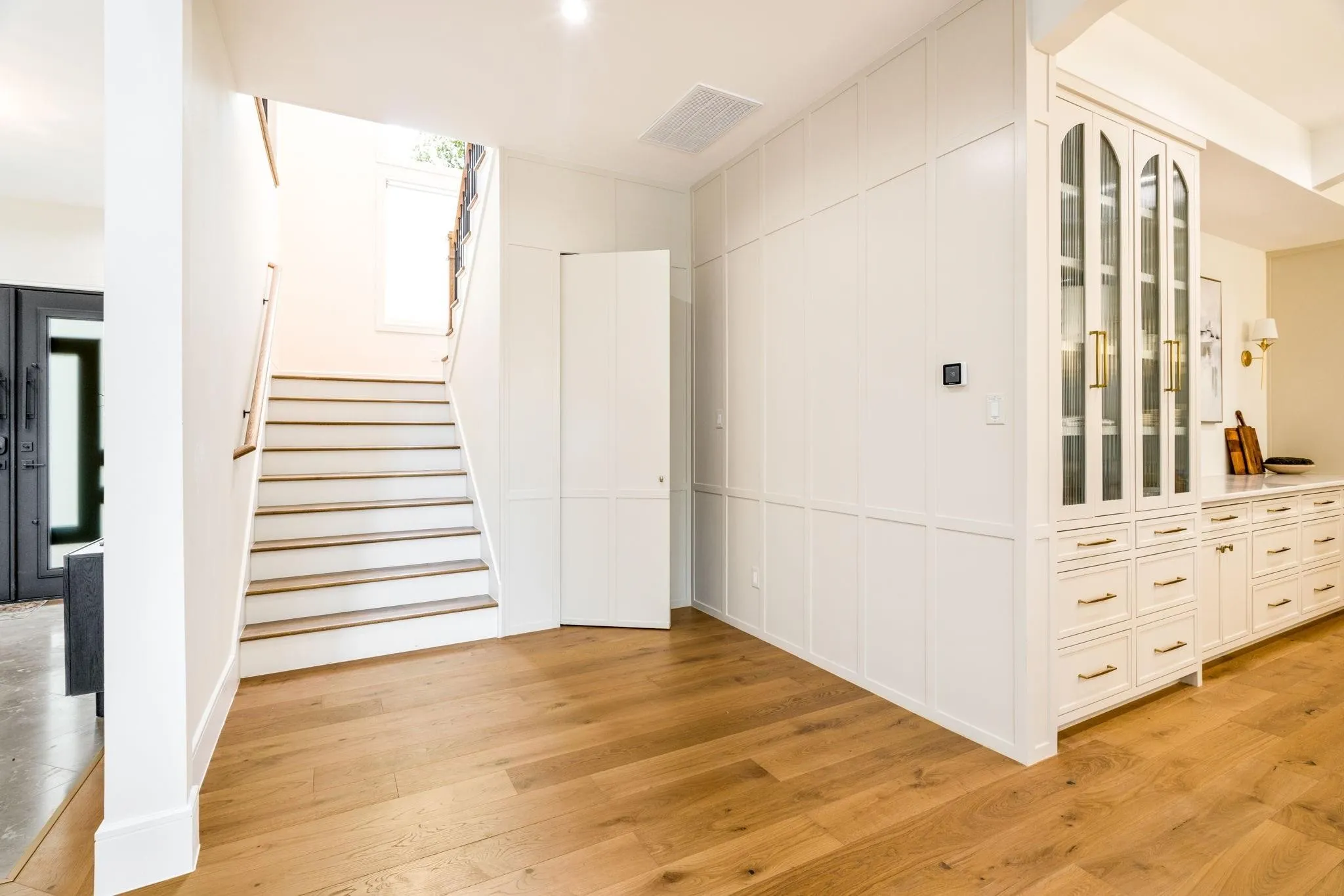 Wide plank wood-finish flooring, staircase with wood-finish treads, custom paneled walls, and built-in cabinetry with glass-front uppers and gold-tone hardware