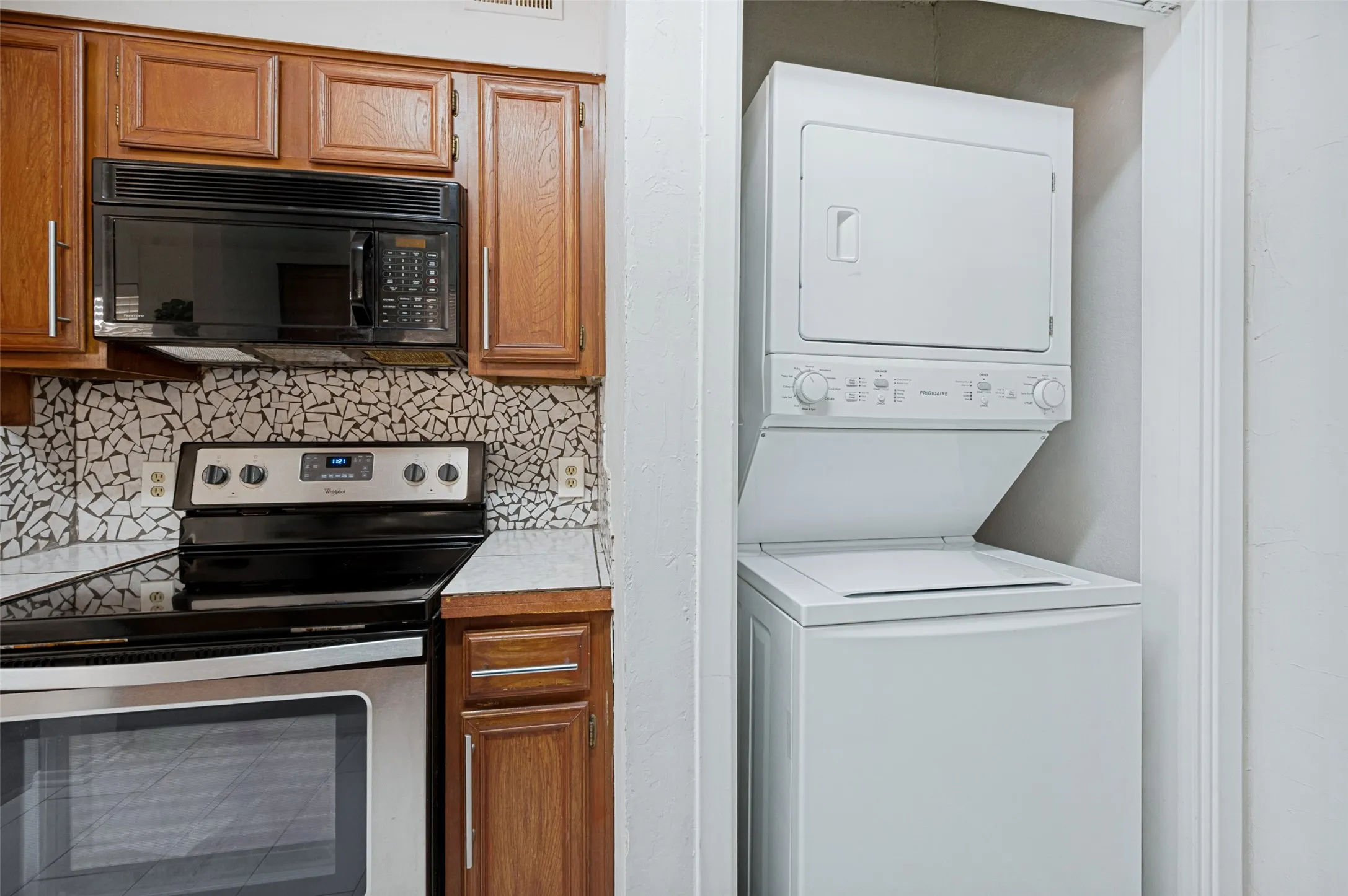 Wood cabinetry, mosaic tile backsplash, stainless steel range with overhead microwave, and a stacked washer/dryer unit