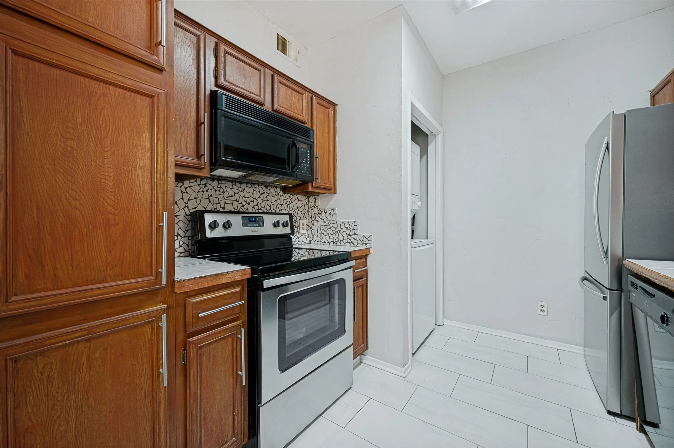 Wood cabinetry, mosaic tile backsplash, black over-the-range microwave, stainless steel range, and light-toned tile flooring