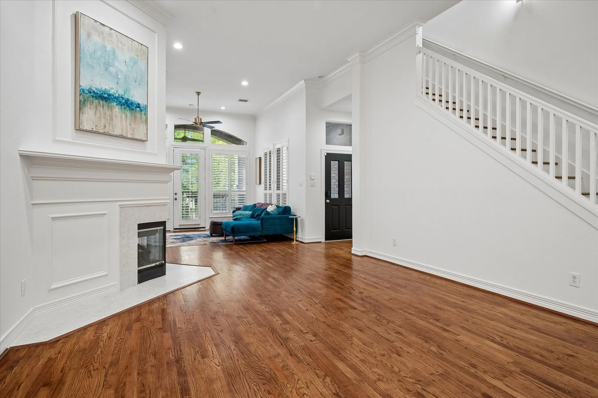 Spacious open-concept living area featuring rich wood-finish flooring, a gas fireplace with white surround and mantel, recessed ceiling lighting, and a ceiling fan