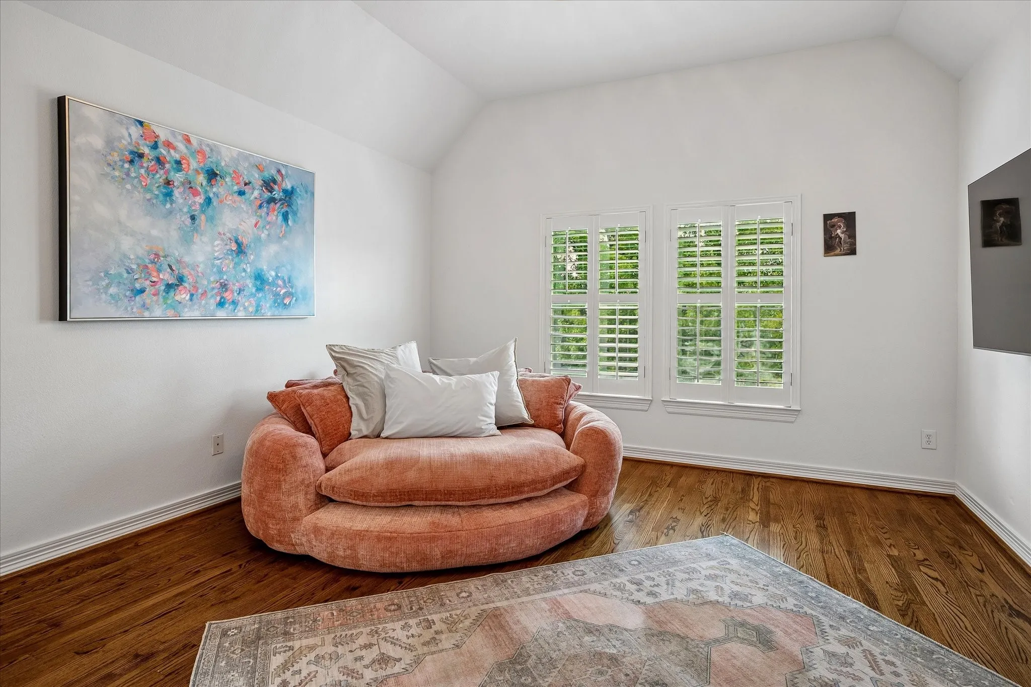 Bright corner room featuring hardwood flooring, plantation shutter-style window treatments with integrated louvers, and an abstract artwork accent wall