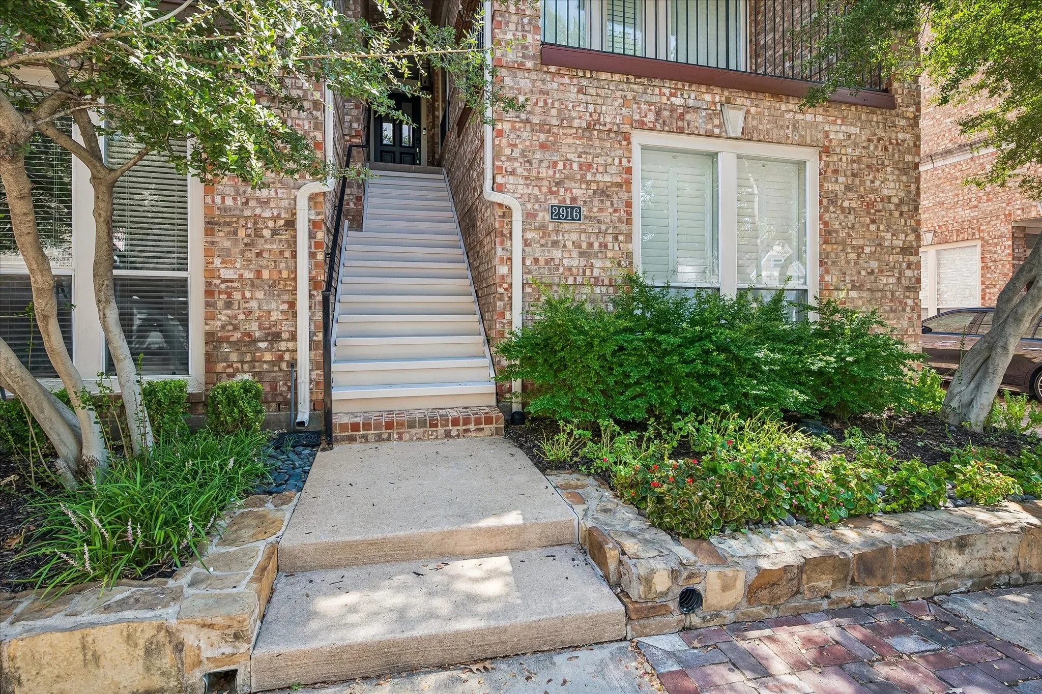 Brick exterior with white window frames, a stone retaining wall, and a concrete pathway leading to an exterior staircase with dark metal railings