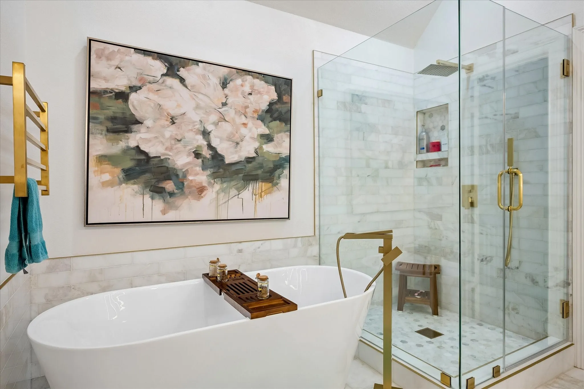 Freestanding soaking tub with a floor-mounted gold-tone faucet, glass-enclosed shower with gold-tone fixtures, marble-look subway tile, and a built-in shower niche