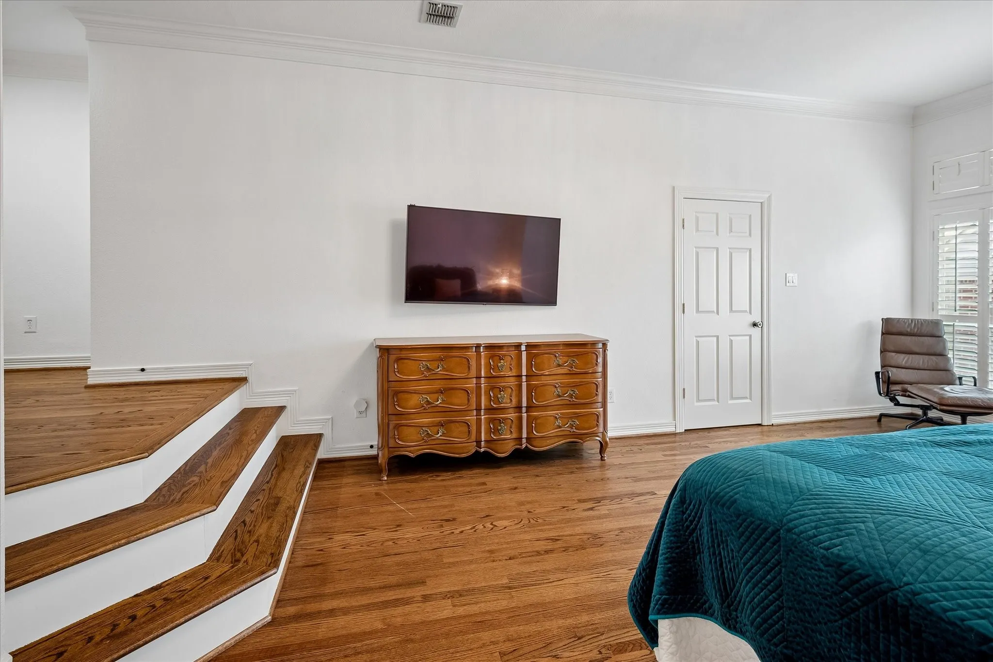 Spacious primary bedroom featuring hardwood flooring, wall-mounted flat-screen television, wooden dresser with multiple drawers, and an ensuite bathroom