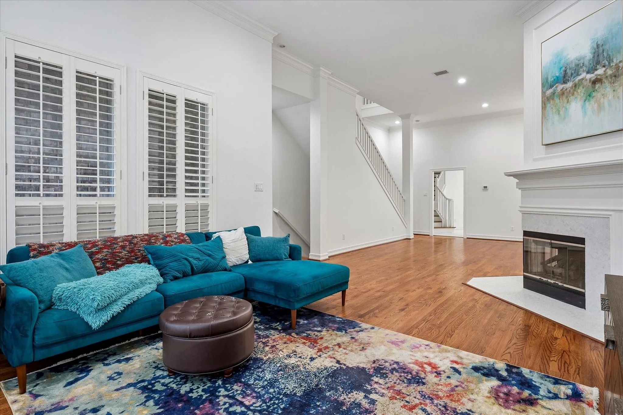 Living area featuring wood-finish flooring, a white-mantled fireplace, staircase with white railing, recessed lighting, and tall windows with plantation shutters