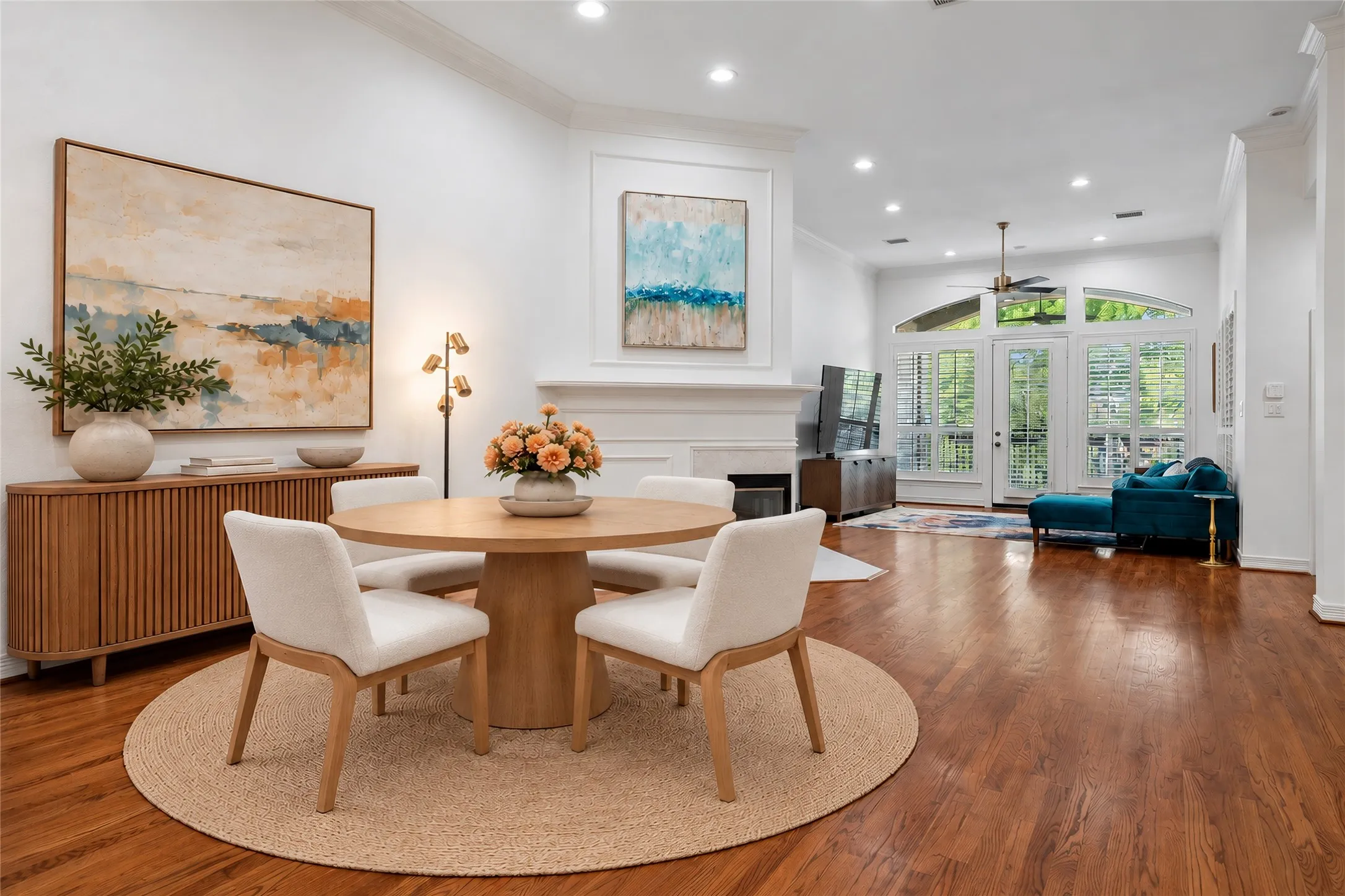 Sophisticated open-concept dining and living area featuring warm wood-finish flooring, a round pedestal dining table, cream upholstered chairs, and recessed ceiling lighting