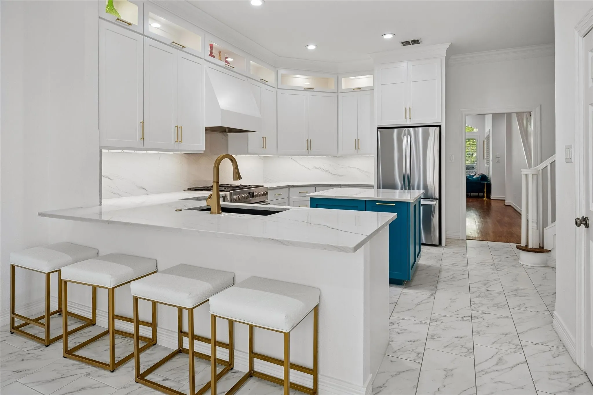 White shaker cabinetry with gold-tone hardware, stainless steel appliances, a center island with a stone countertop, a gold-tone gooseneck faucet, and white marble-look flooring