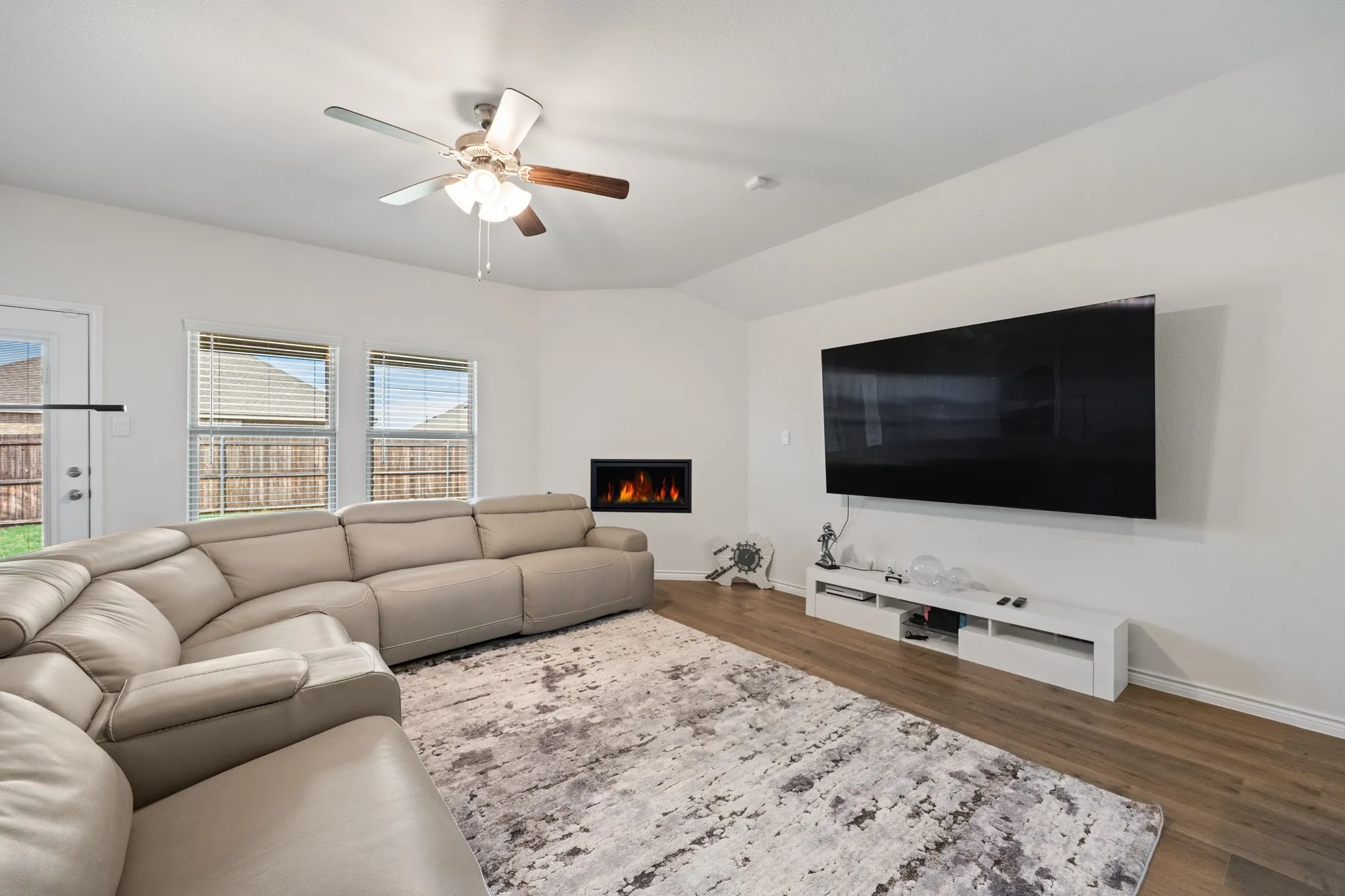 Wood-finish flooring throughout, ceiling fan with light fixture, built-in electric fireplace, and multiple windows with blinds