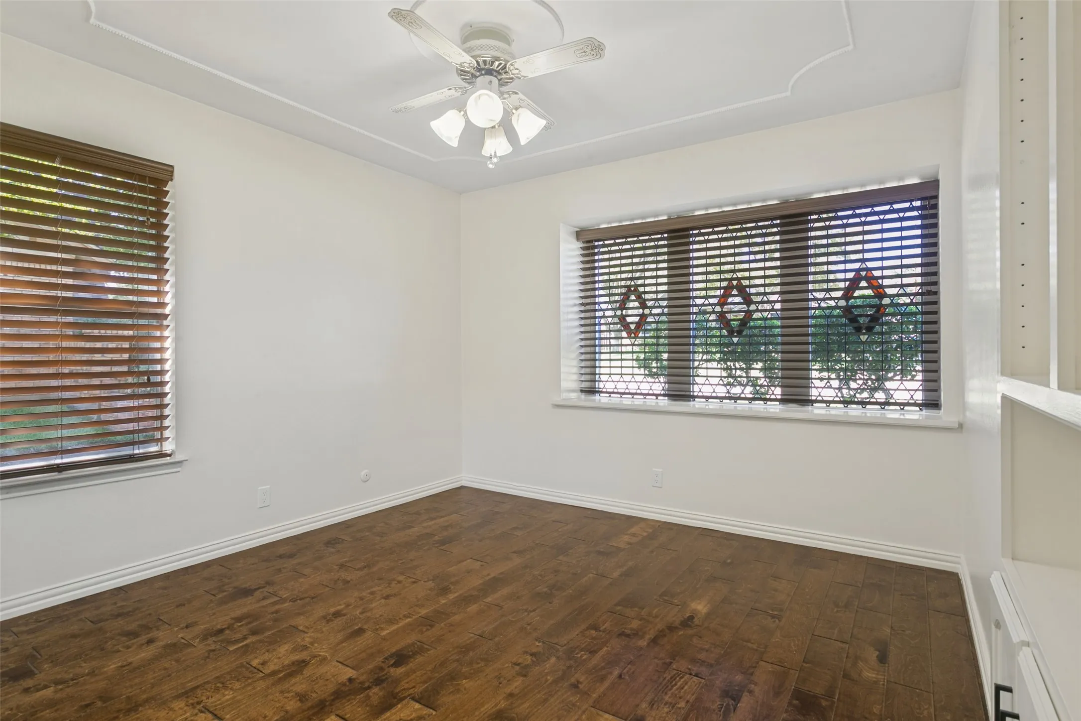 Wood-finish flooring, white walls, decorative window blinds, ceiling fan with integrated lighting, and a built-in shelving unit