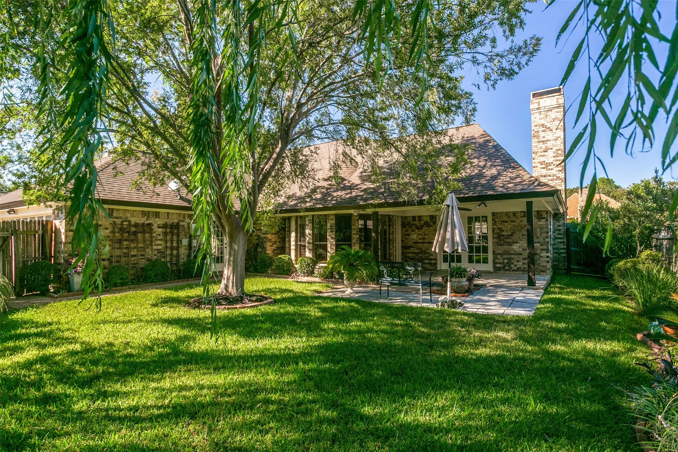 Expansive backyard featuring a stone patio, covered porch with columns, mature trees, and a brick exterior