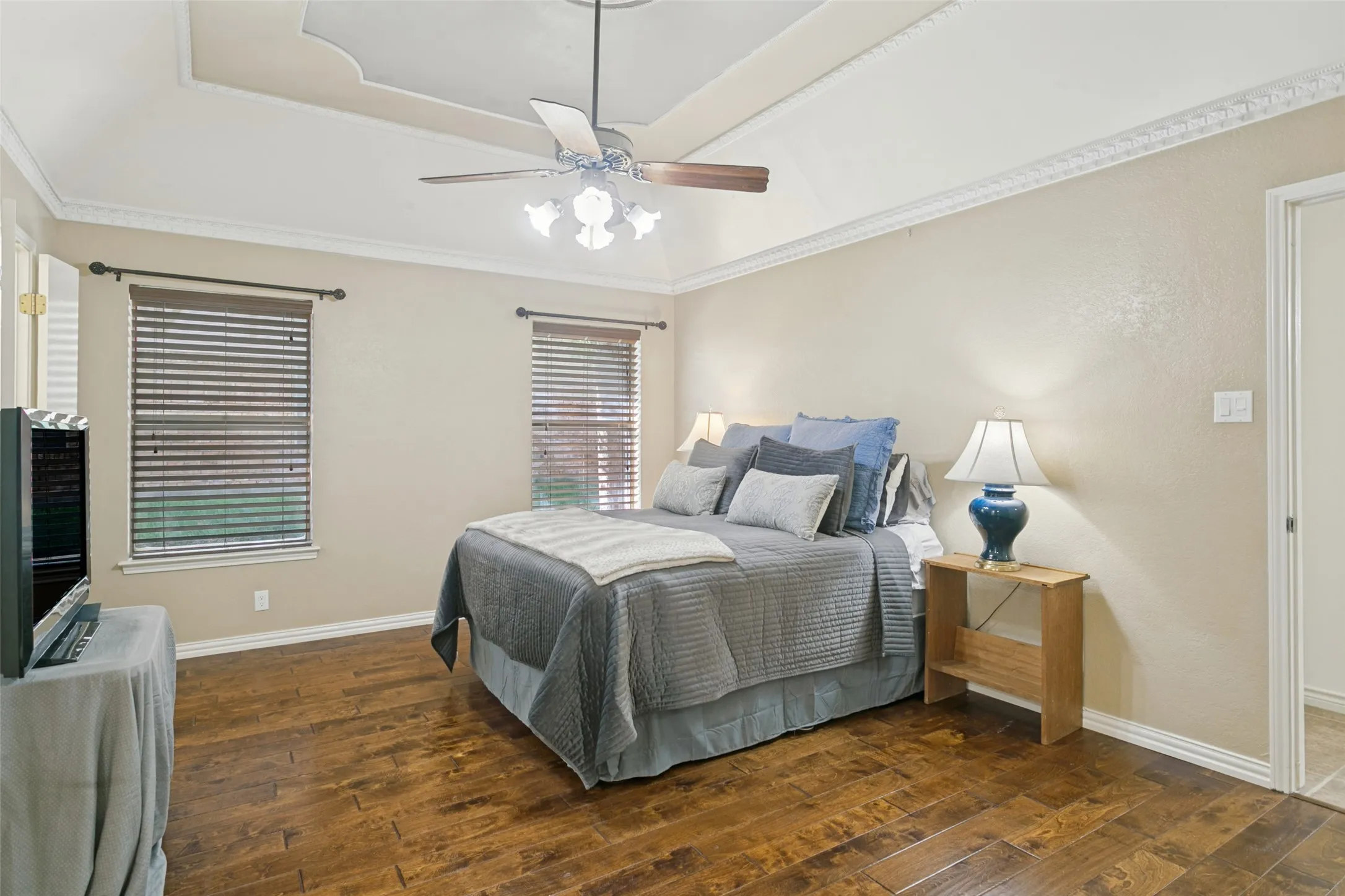 Wood-finish flooring, tray ceiling, crown molding, two windows with blinds, and a ceiling fan with integrated lighting