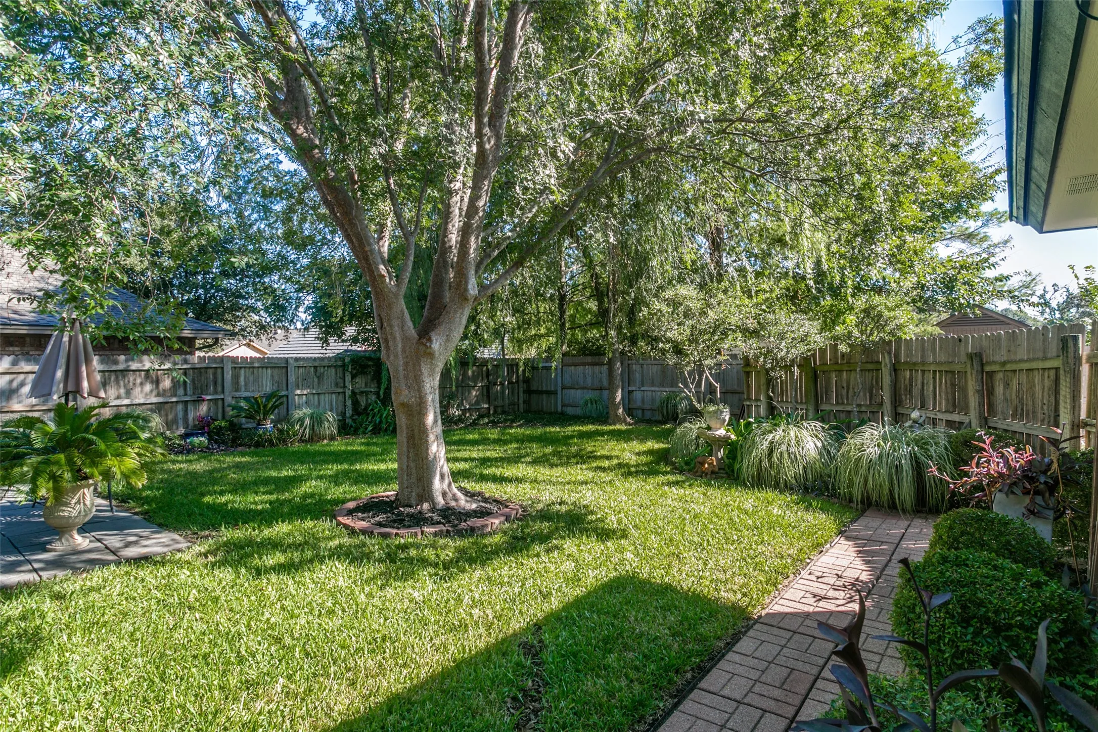 Expansive backyard featuring a manicured lawn, mature trees, a brick paver pathway, and a wood privacy fence