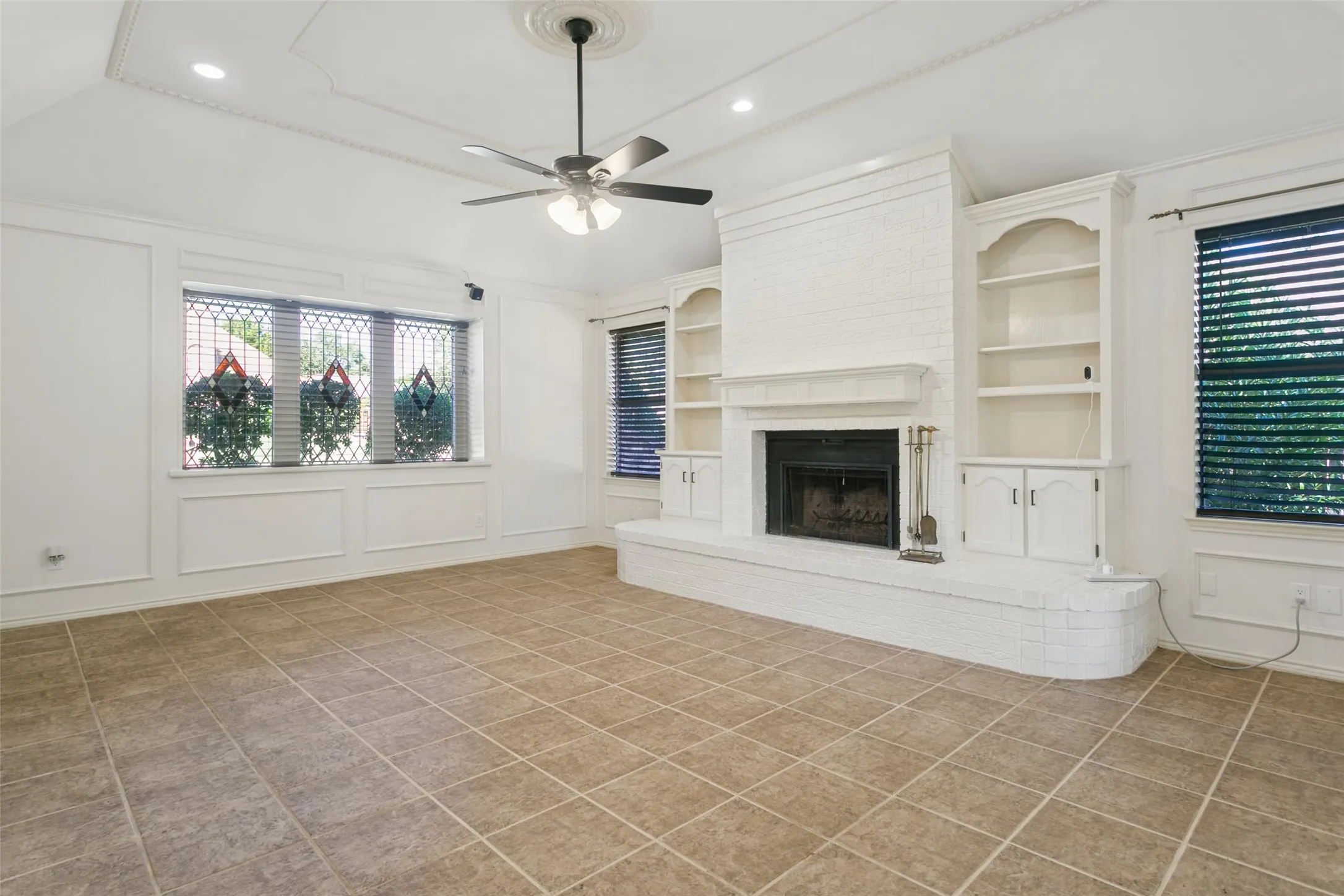 Spacious room featuring tile flooring, a white brick fireplace, built-in shelving, a tray ceiling, and a ceiling fan