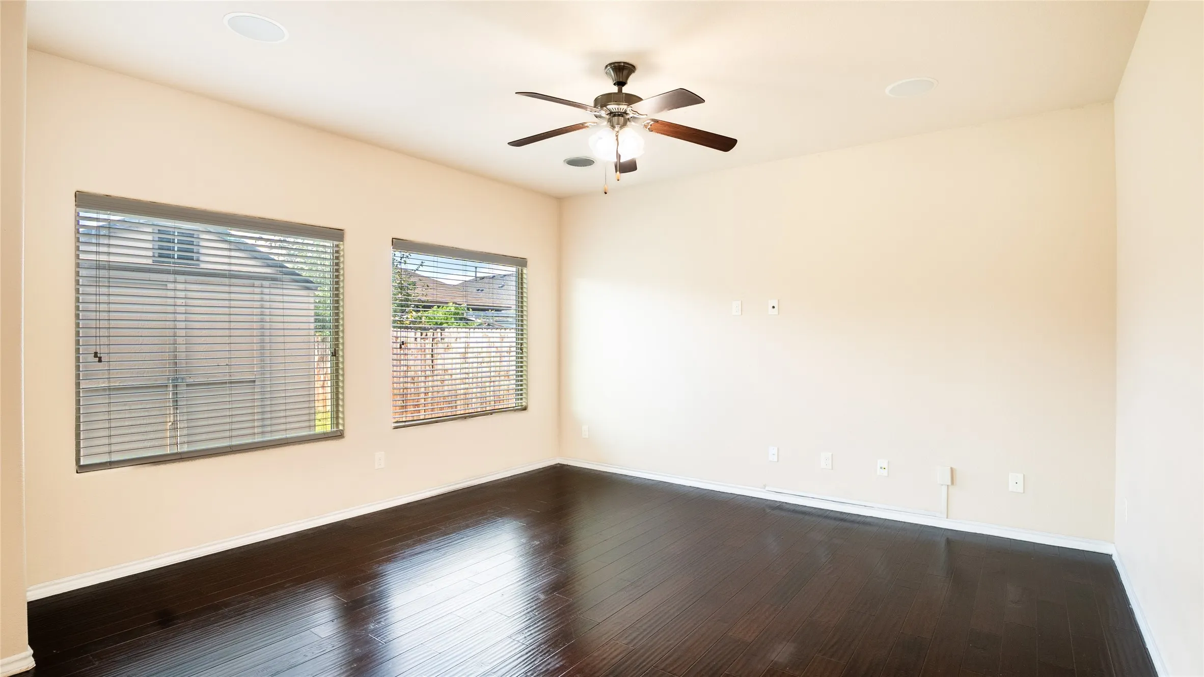 Dark wood-finish flooring, two windows with blinds, ceiling fan with integrated lighting, and recessed ceiling speakers