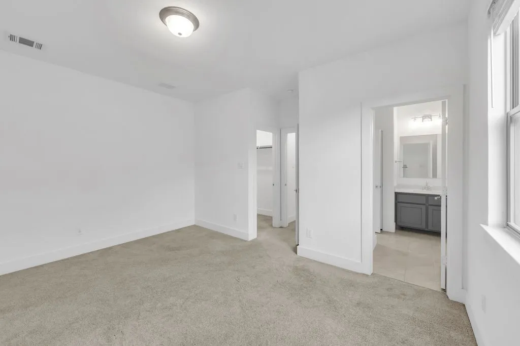 Carpeted flooring, white walls, ceiling-mounted light fixture, window with white trim, and a bathroom with a vanity and mirror