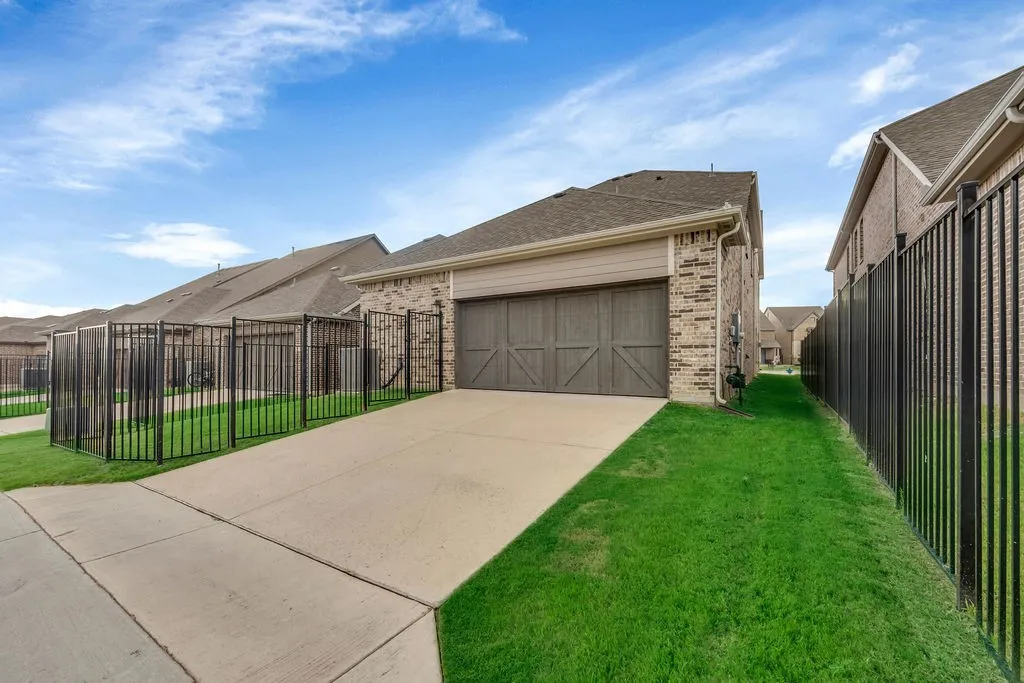 Concrete driveway with a wood-finish garage door, brick exterior, and a black metal fence enclosing a grass lawn