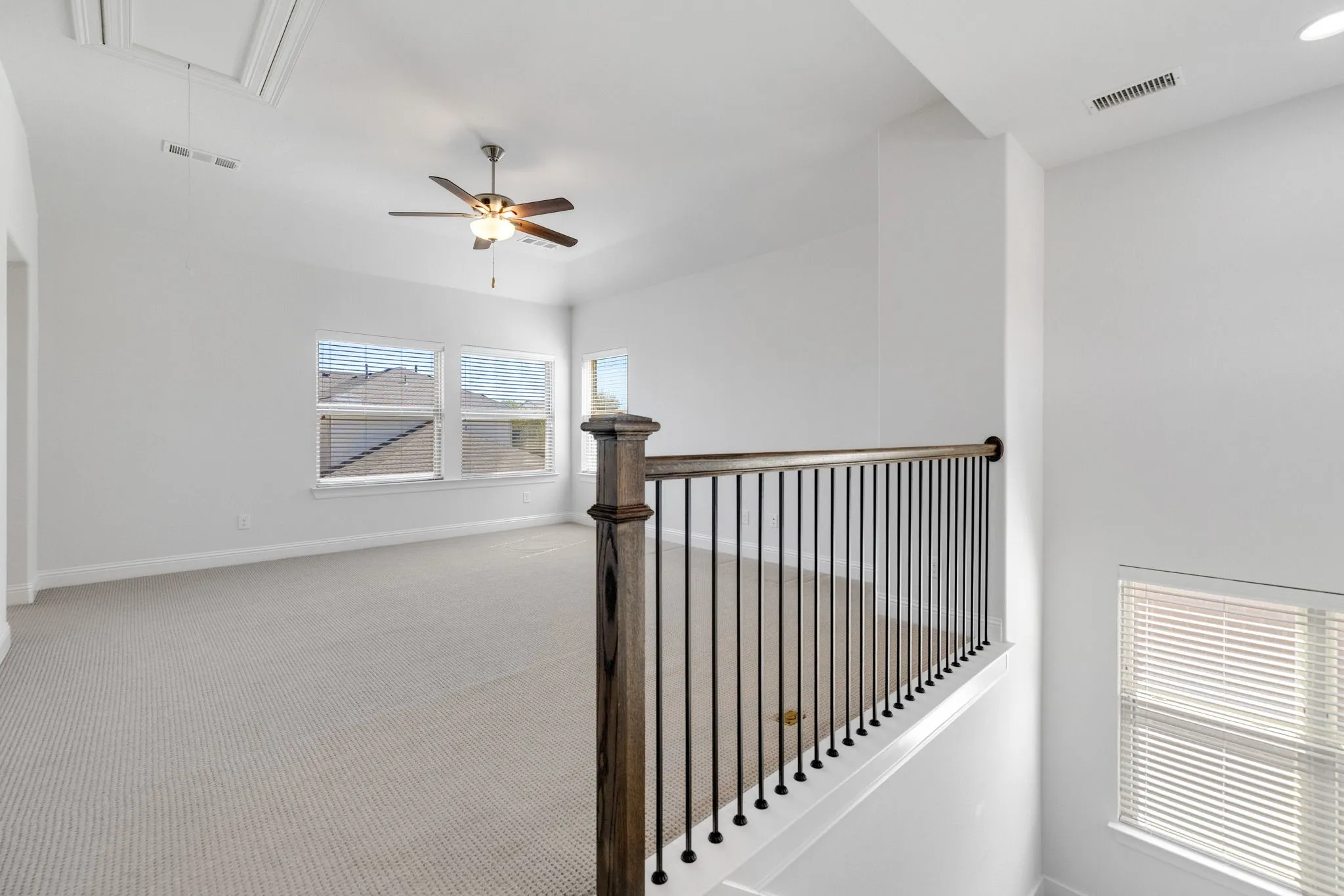 Spacious second-level area featuring light-colored carpeting, multiple windows with blinds, a ceiling fan with integrated lighting, and a stair railing with wood newel posts and black metal balusters