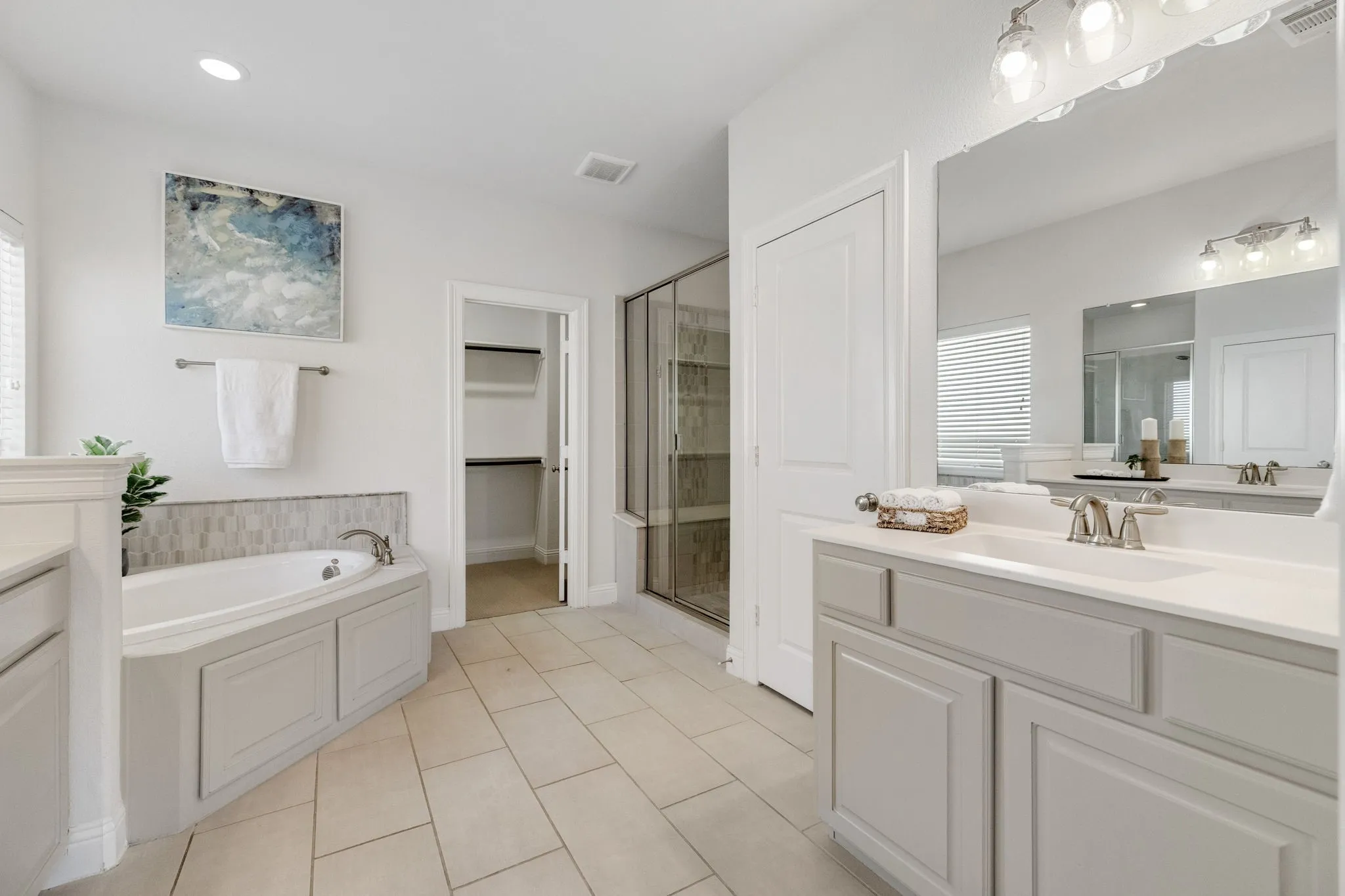 Spacious bathroom with neutral tile flooring, a built-in corner soaking tub, a separate glass-enclosed shower, a large vanity with an integrated sink, and an expansive wall-mounted mirror