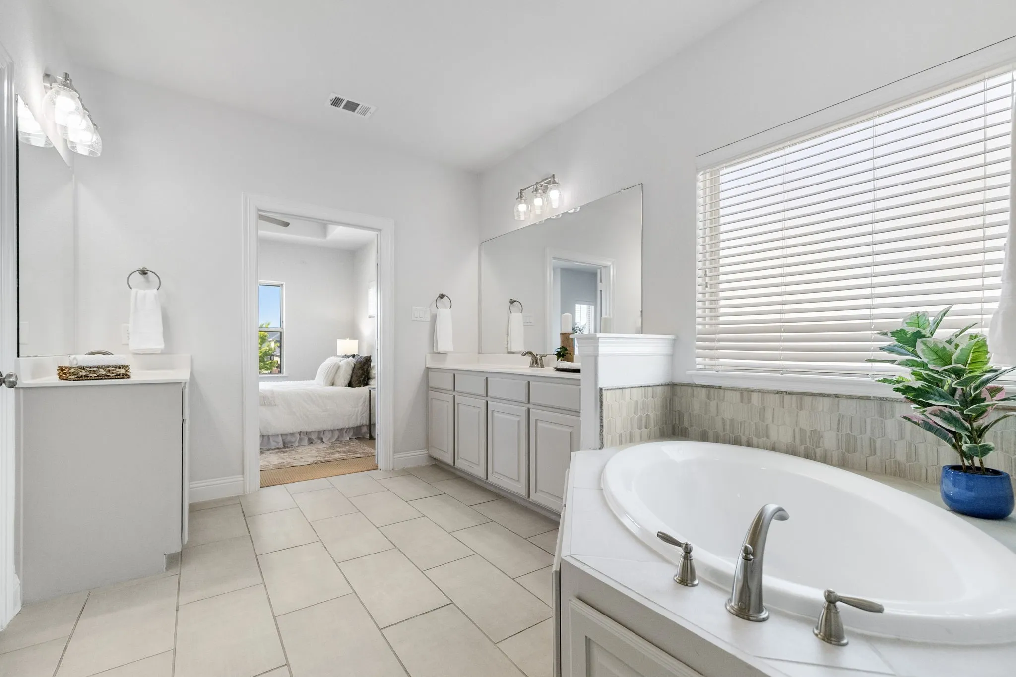 Built-in soaking tub with brushed nickel fixtures, dual vanities with white countertops, tile flooring, and a large window with blinds