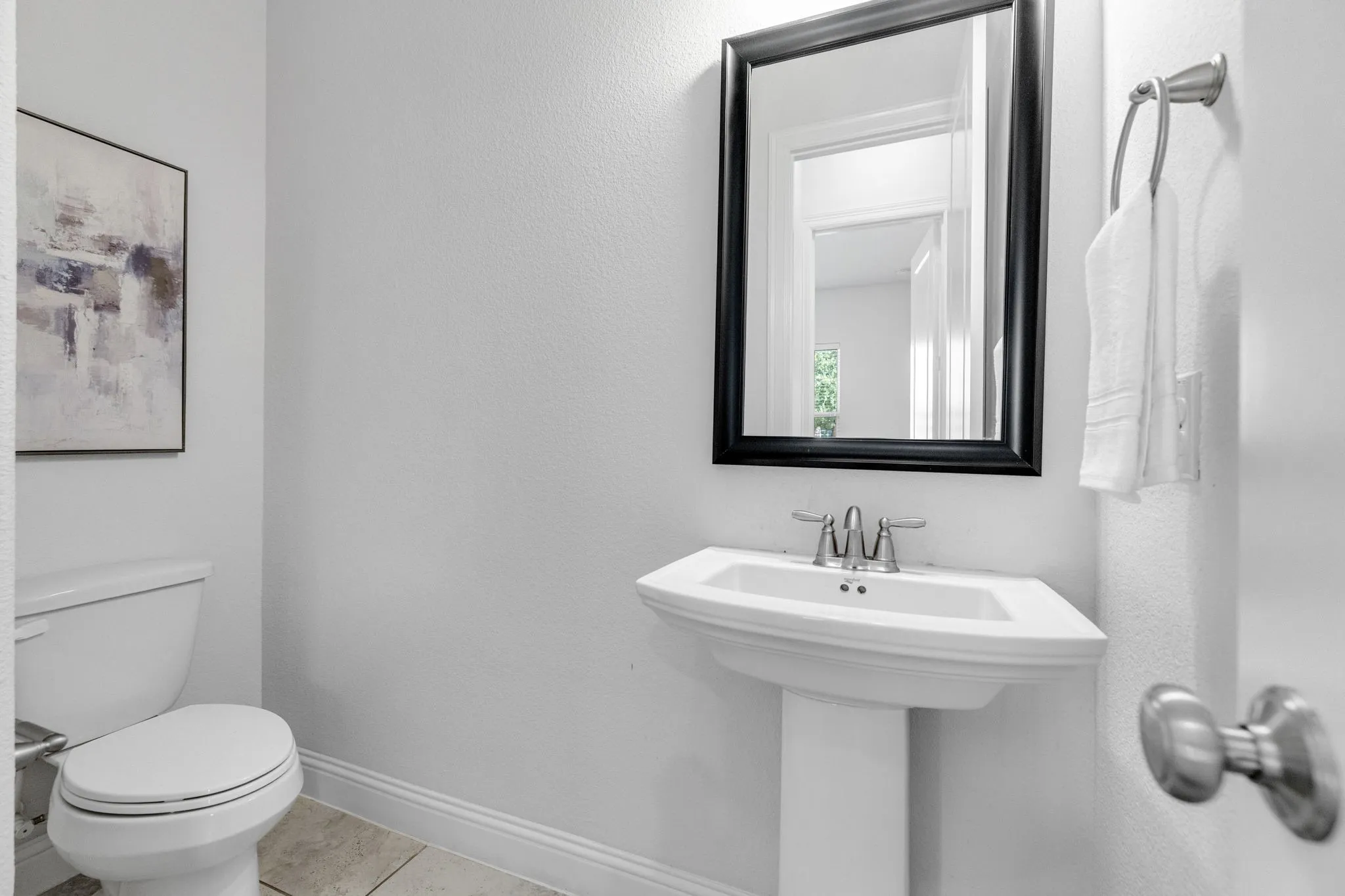 Elegant half bath featuring a pedestal sink with polished chrome faucet, framed mirror with black trim, ceramic tile flooring, and clean white wall finishes