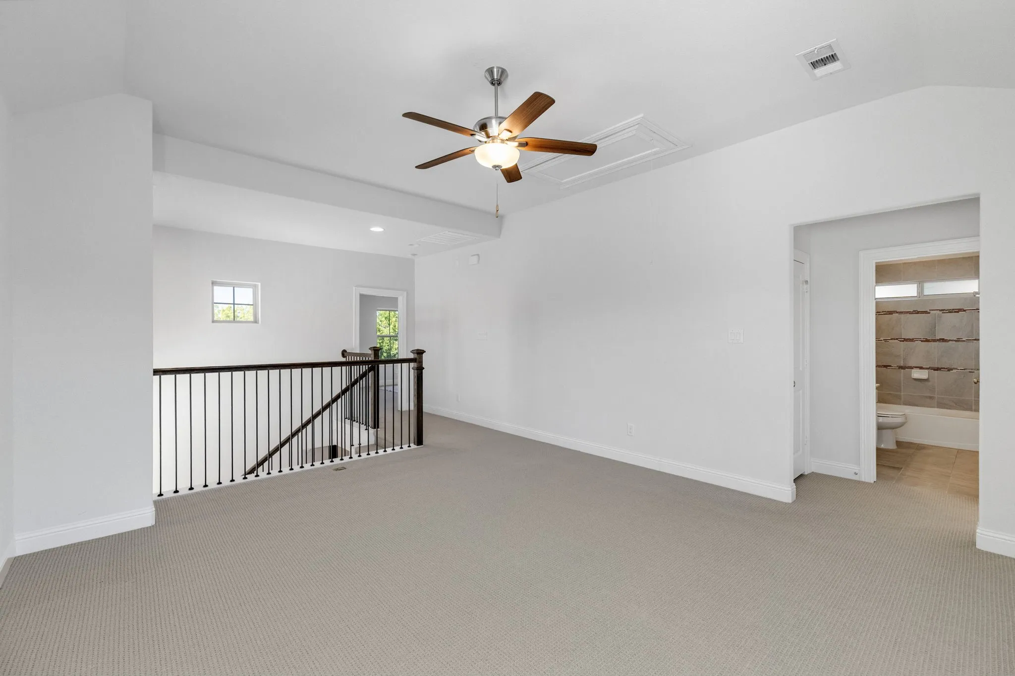 Carpeted loft area with a ceiling fan, wrought iron and wood railing, and a window