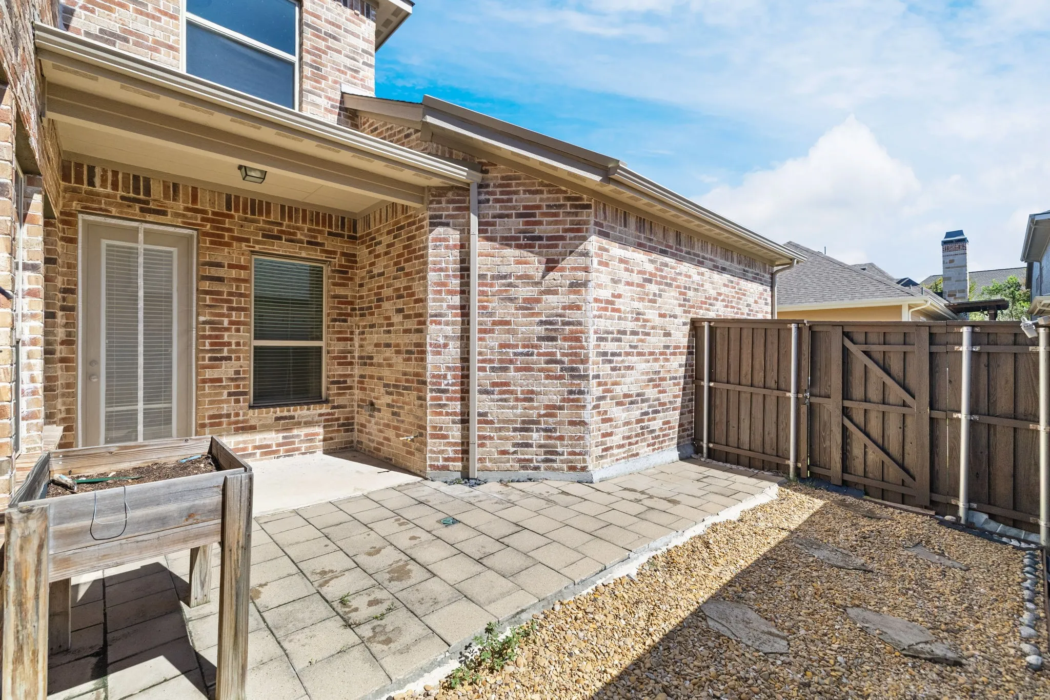 Brick exterior with a covered patio, paved hardscaping, a privacy fence, and a gravel-covered area