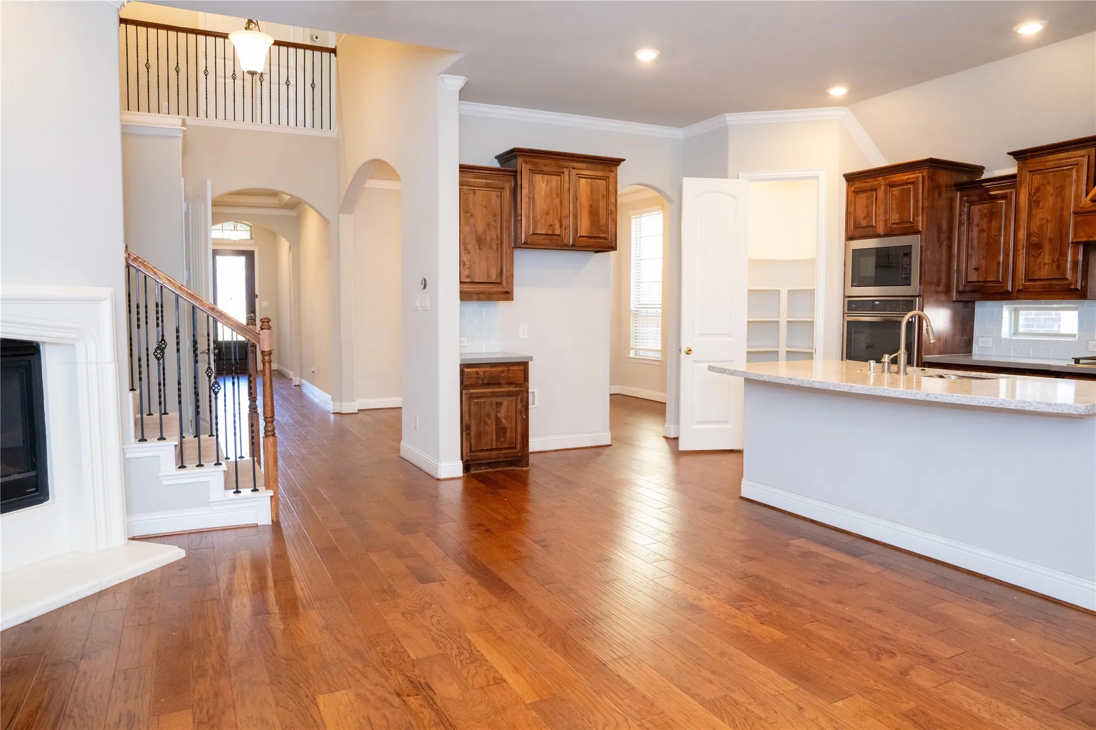 Hardwood flooring, arched doorways, built-in kitchen appliances, kitchen island with sink, white shaker cabinetry with dark wood-finish cabinetry