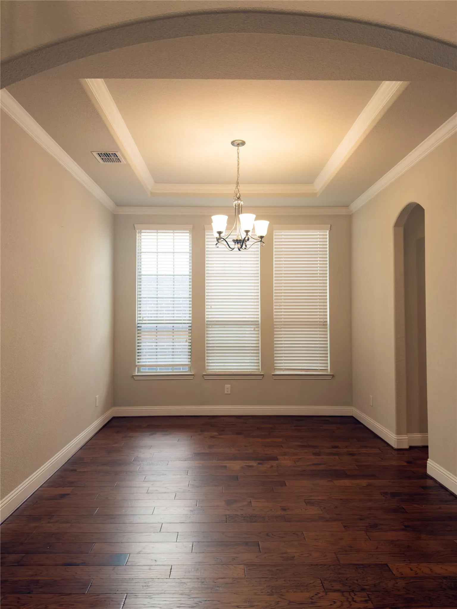 Recessed ceiling with layered trim, chandelier lighting, three windows with blinds, wood-finish flooring, and arched doorway