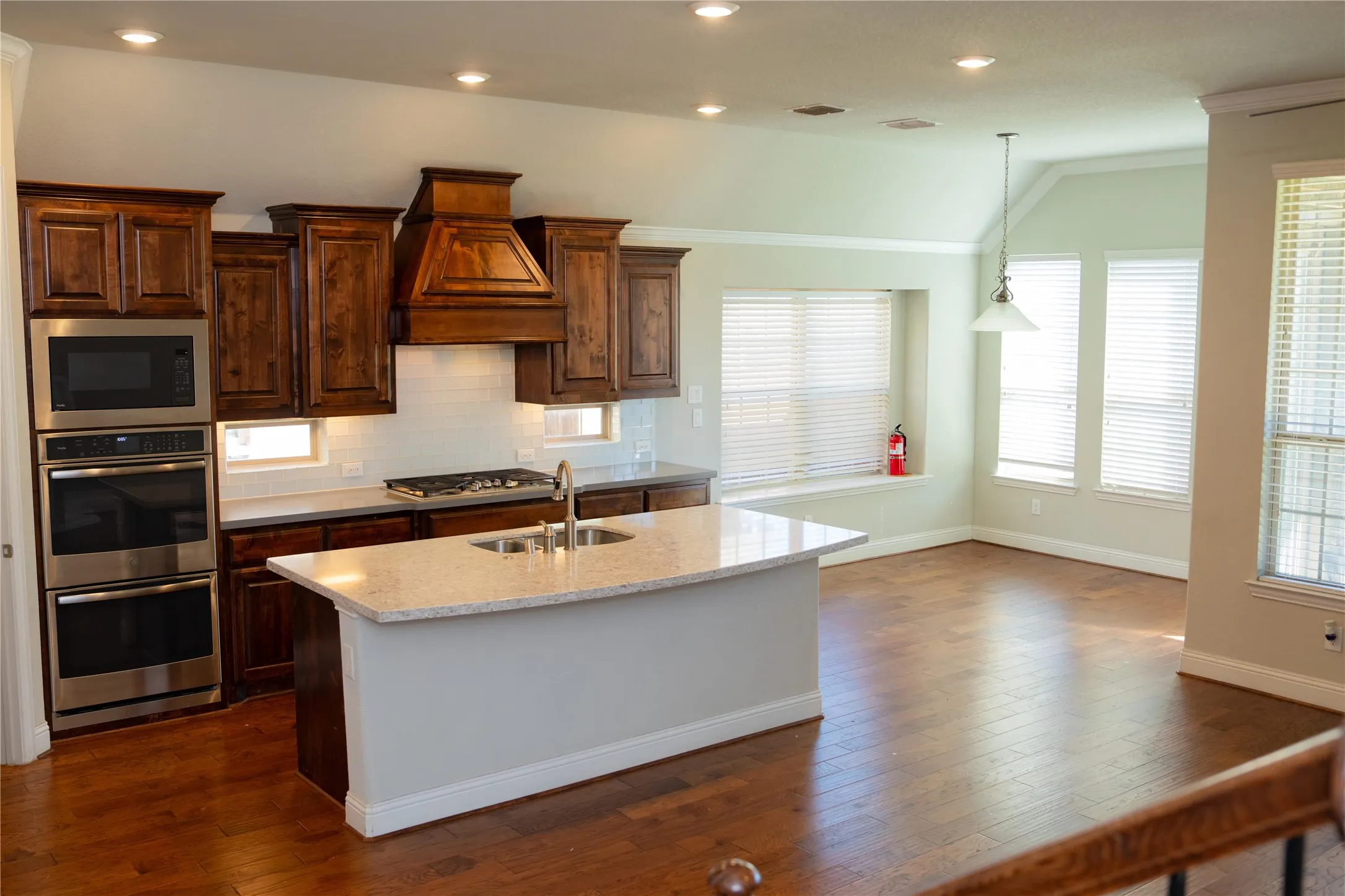 Wood-finish flooring throughout, kitchen with wood cabinetry and stainless steel double ovens, central island with an undermount sink and light-toned countertop, and recessed lighting