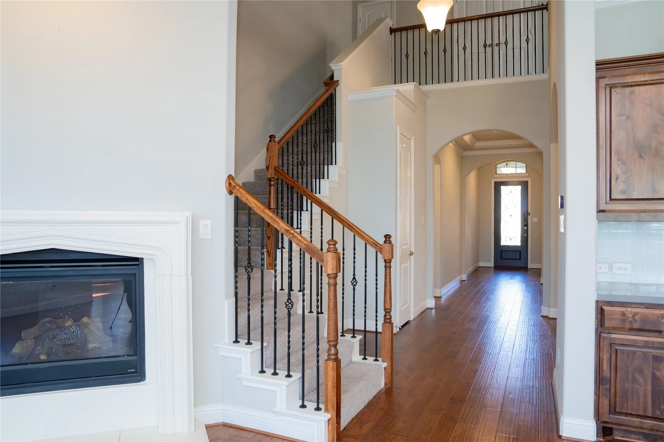 Hardwood flooring, fireplace with white mantel, carpeted staircase with wood handrails and wrought iron balusters, arched entryway, and built-in cabinetry
