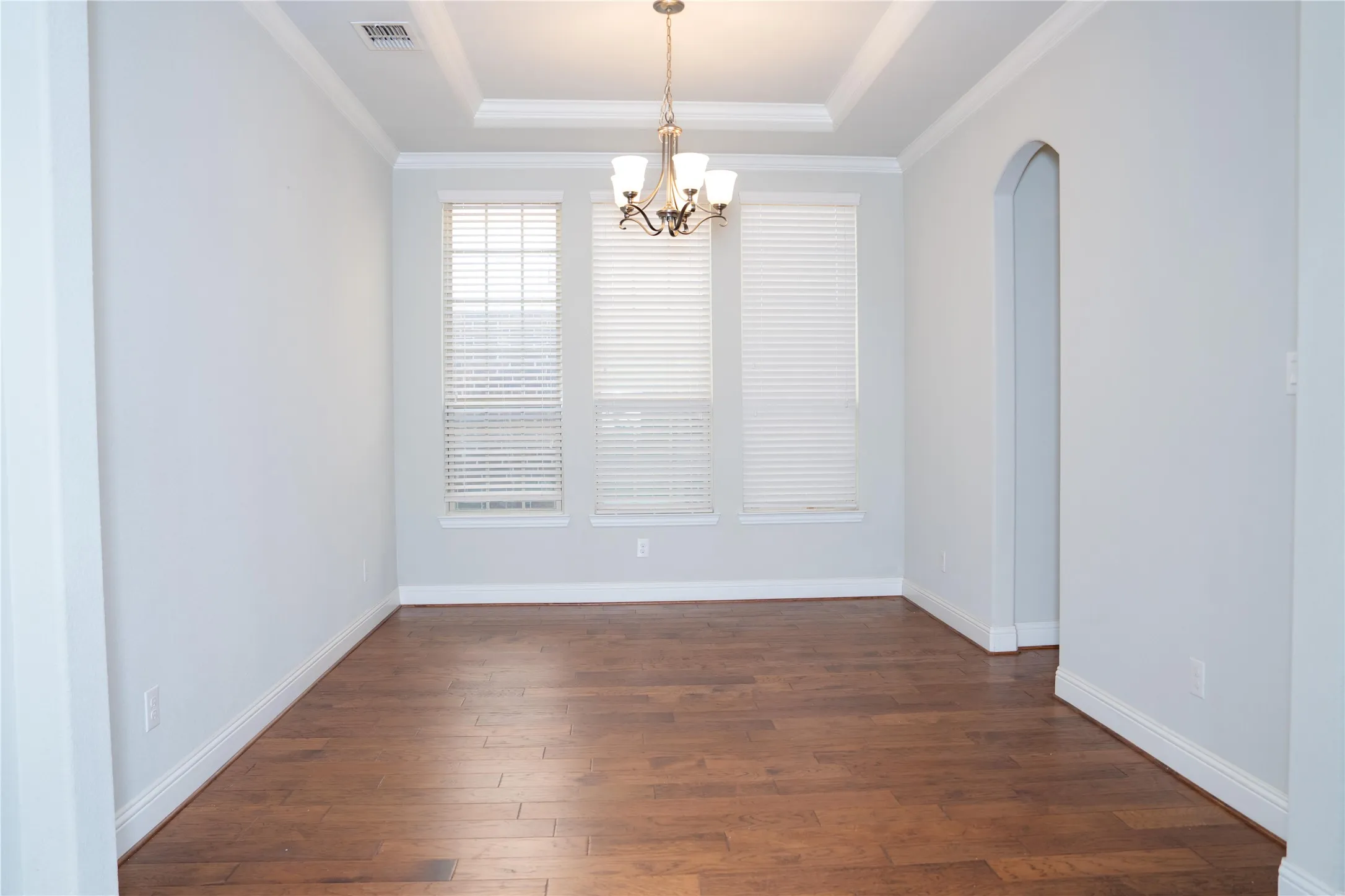 Recessed ceiling with a central chandelier, wood-finish flooring, three large windows with blinds, and an arched doorway