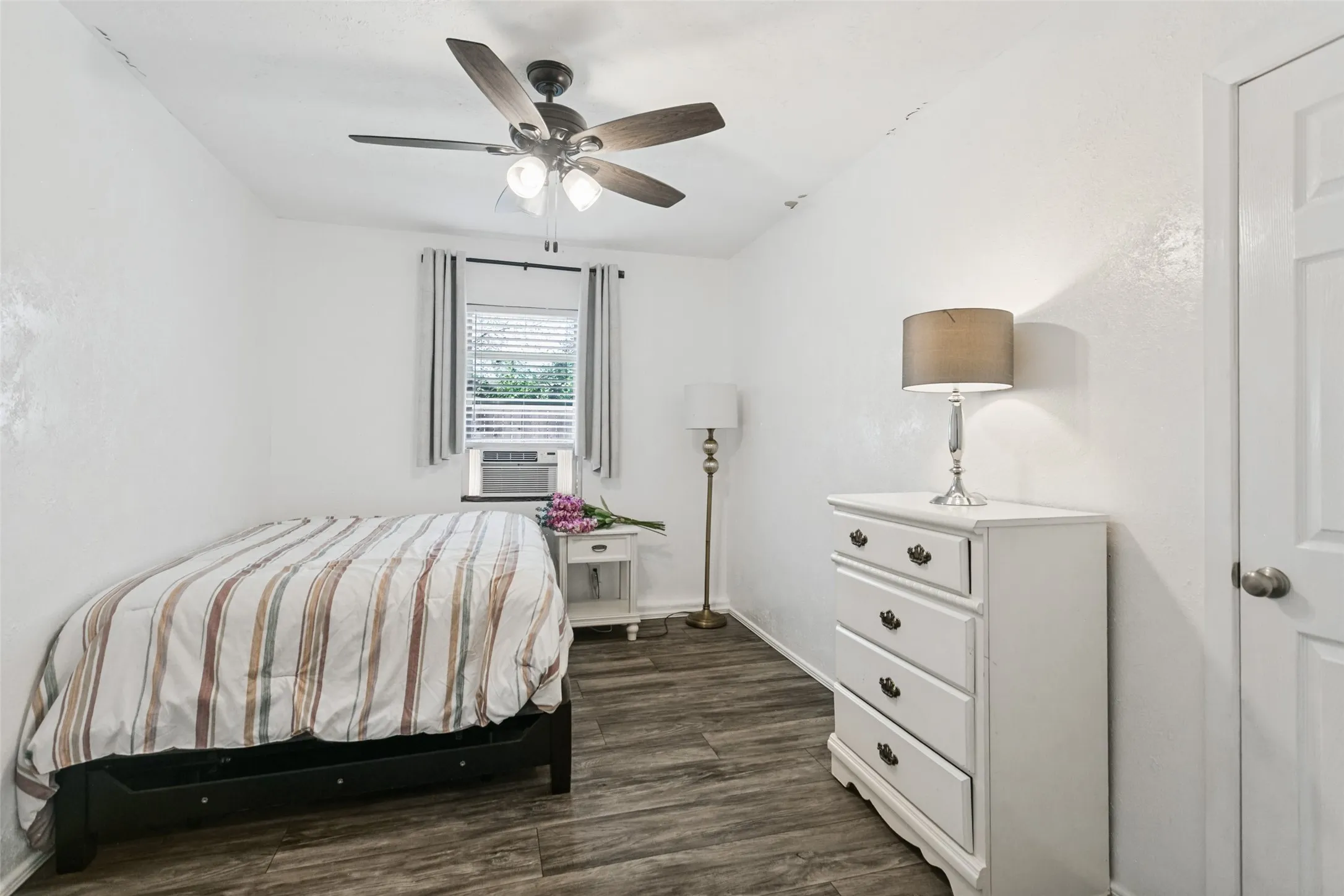 Bright secondary bedroom featuring wood-finish flooring, ceiling fan with integrated lighting, window with air conditioning unit, and white painted cabinetry with multiple drawers for storage