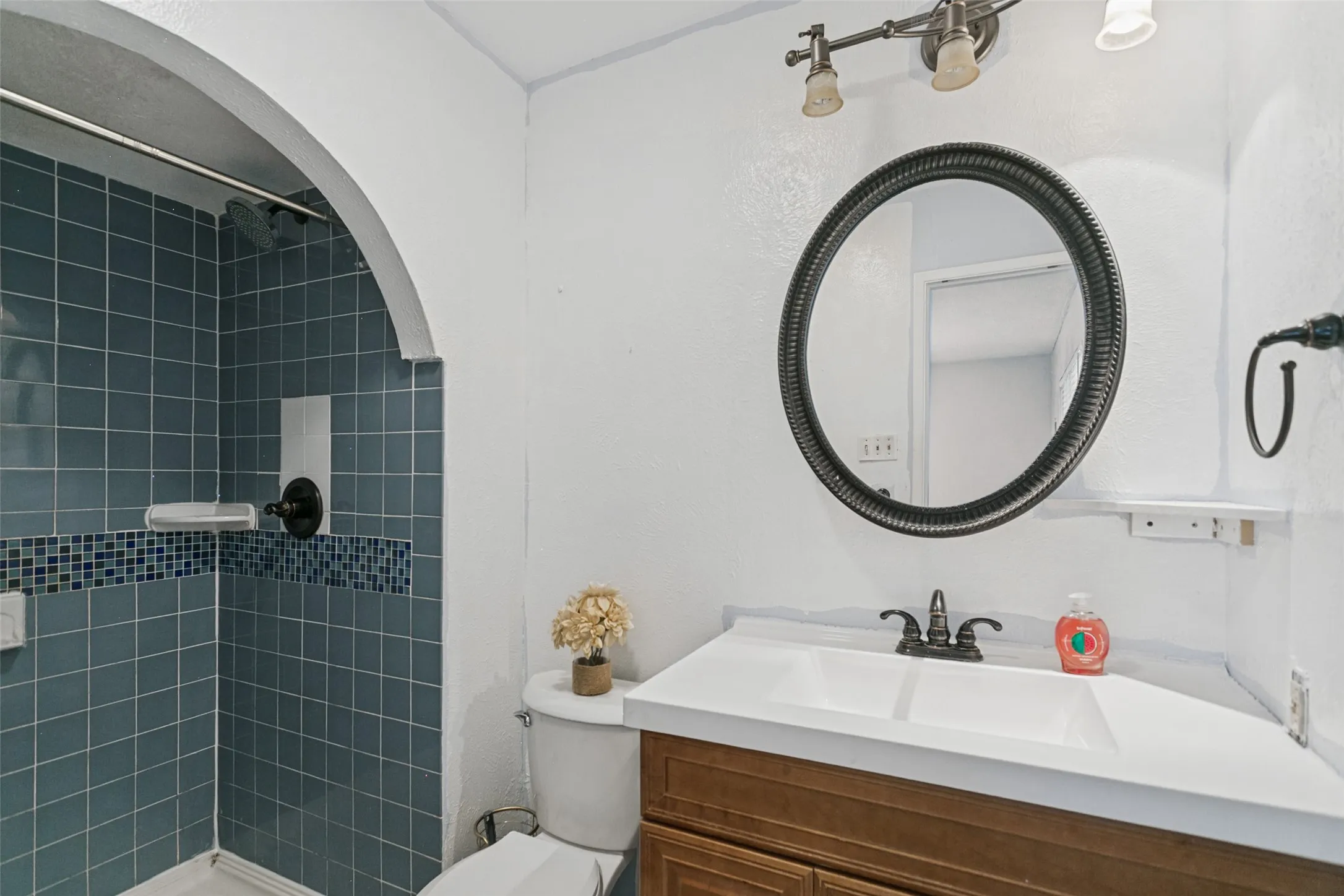 Bathroom featuring an arched shower entrance, blue tile shower surround with mosaic accent, wood-finish vanity with dual sinks, and an oval mirror
