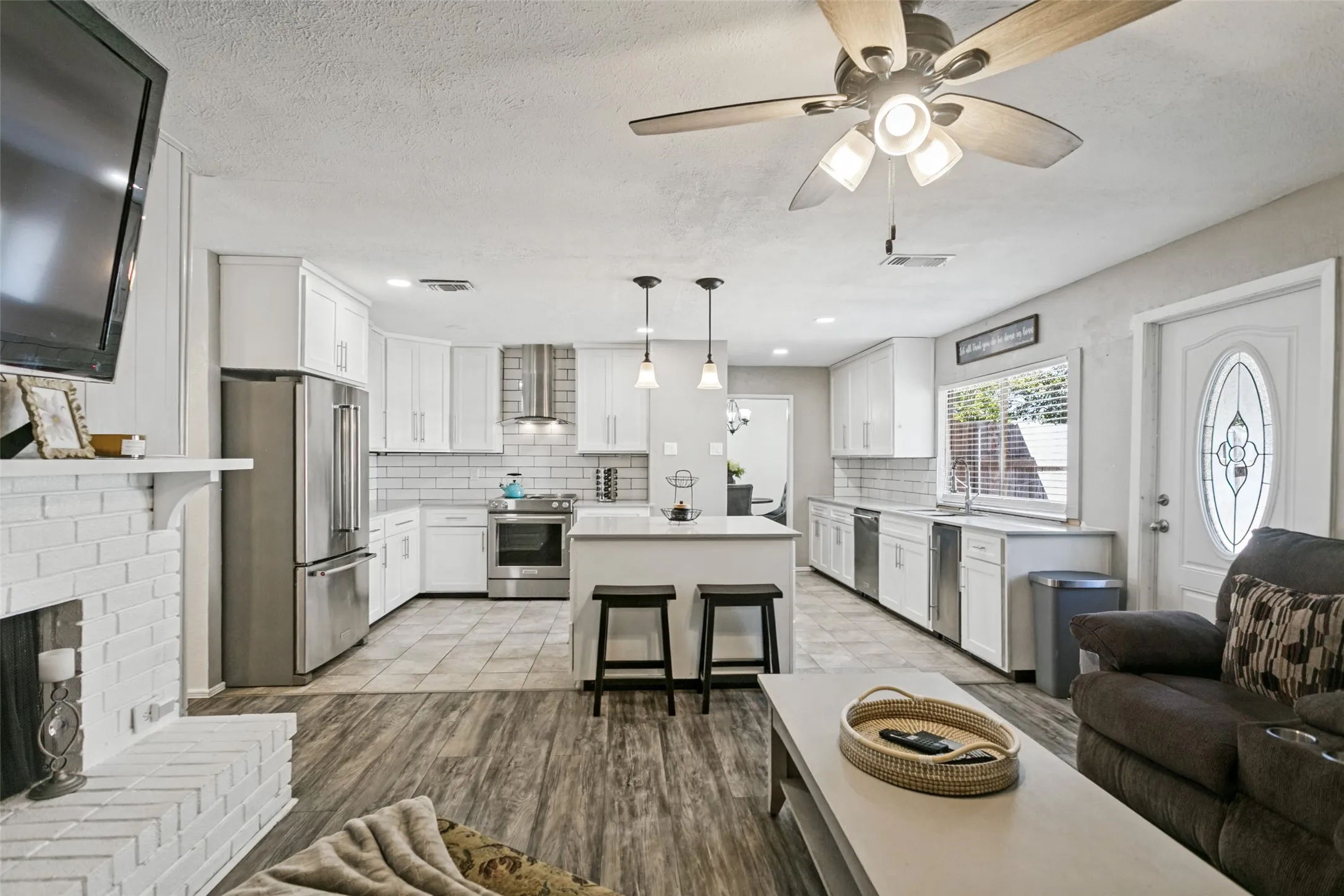 White brick fireplace, wood-finish flooring, white kitchen cabinetry, stainless steel appliances, and a kitchen island with pendant lighting