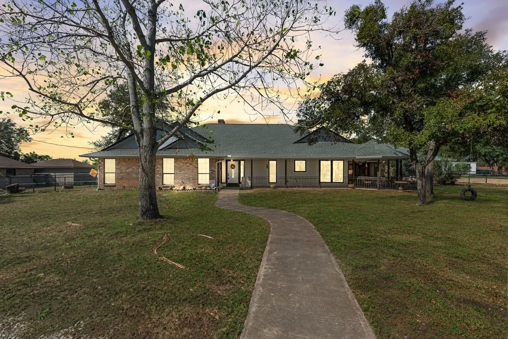 Brick and siding exterior with a green shingle roof, featuring a covered porch, a concrete walkway, and established trees