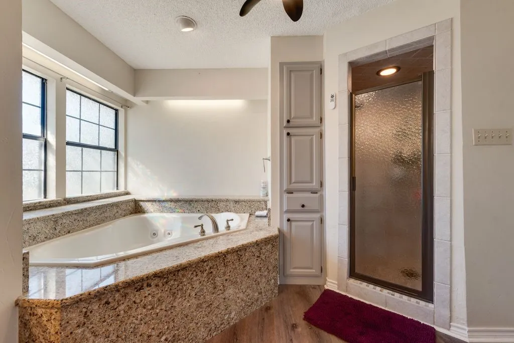 Built-in jetted tub with a granite surround, adjacent windows, a separate shower with a textured glass door, and integrated cabinetry