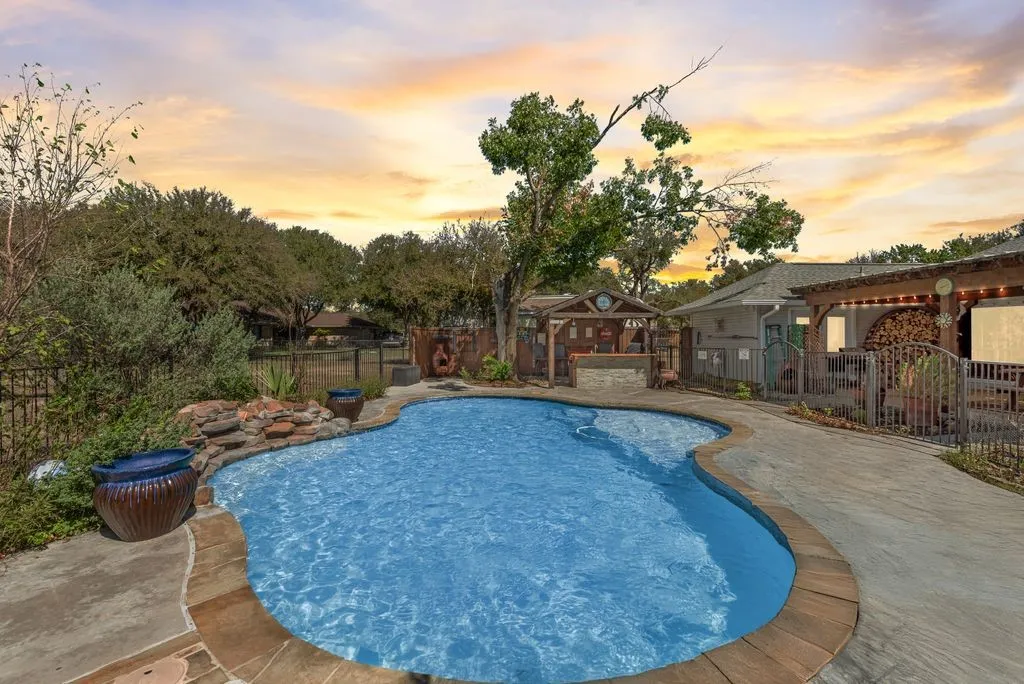 Freeform swimming pool with stone coping, concrete decking, rock waterfall feature, and a covered outdoor kitchen