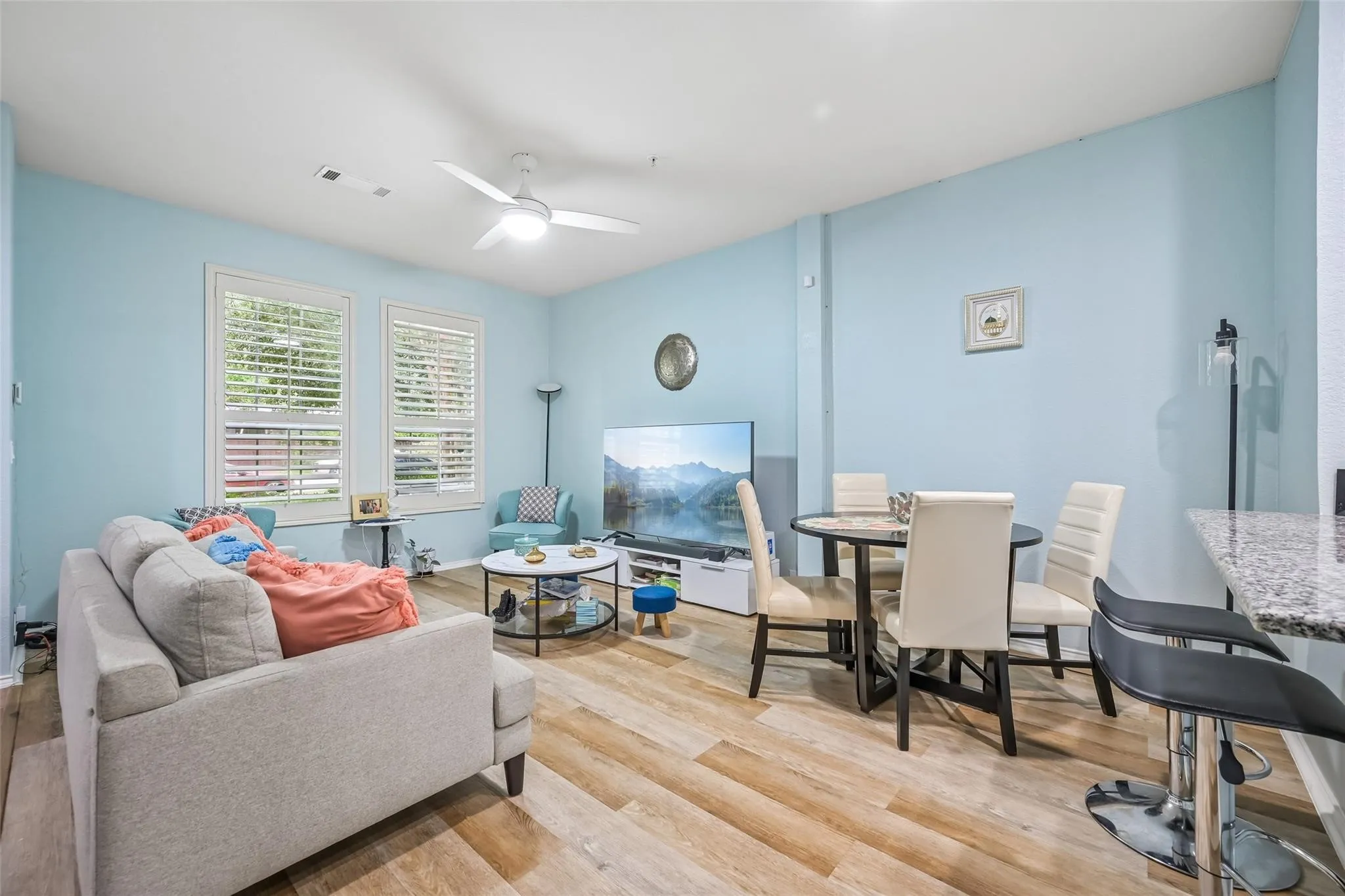 Open-concept living space featuring wood-finish flooring, a ceiling fan with integrated lighting, and multiple windows with plantation shutters