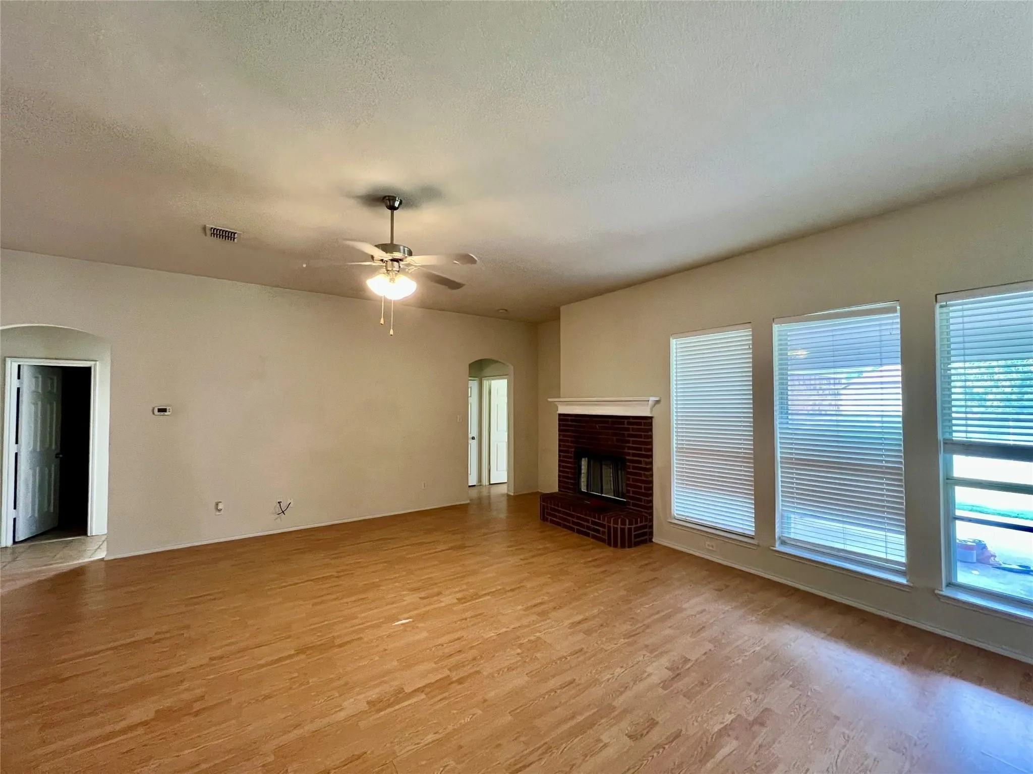 Spacious room featuring wood-finish flooring, a ceiling fan with integrated lighting, a brick fireplace with a white mantel, and multiple windows