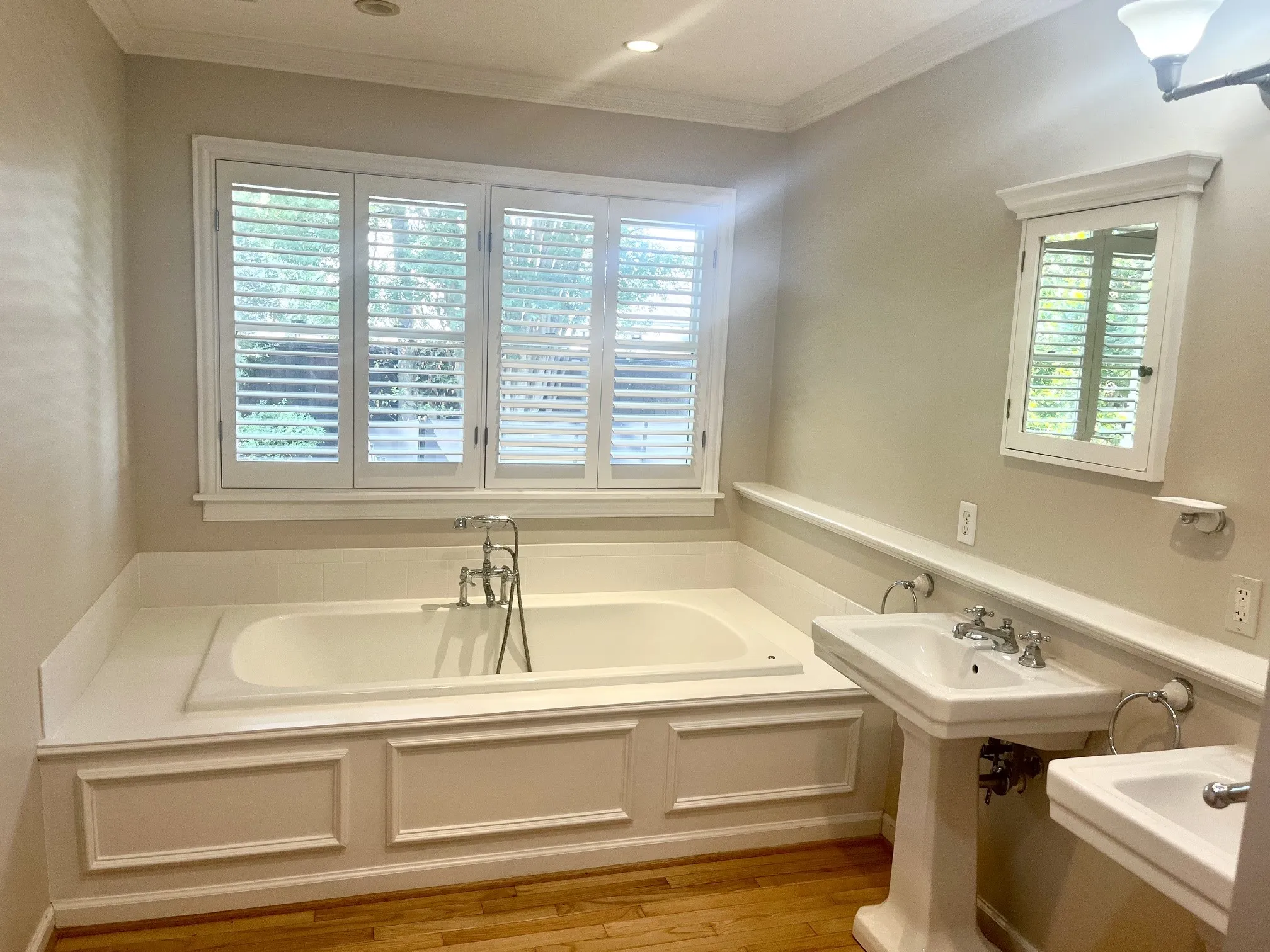 Built-in soaking tub with chrome fixtures, three panel window with plantation shutters, two pedestal sinks, built-in medicine cabinet, and wood-finish flooring