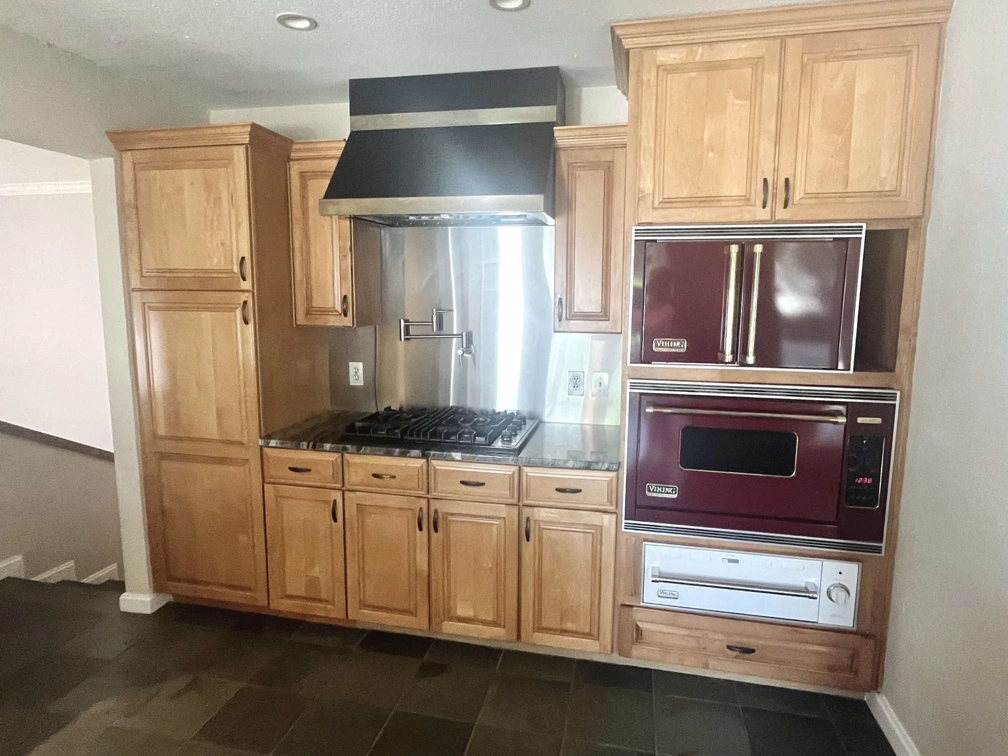Built-in gas cooktop with a stainless steel backsplash, integrated range hood, wood-finish cabinetry, and dark tiled flooring