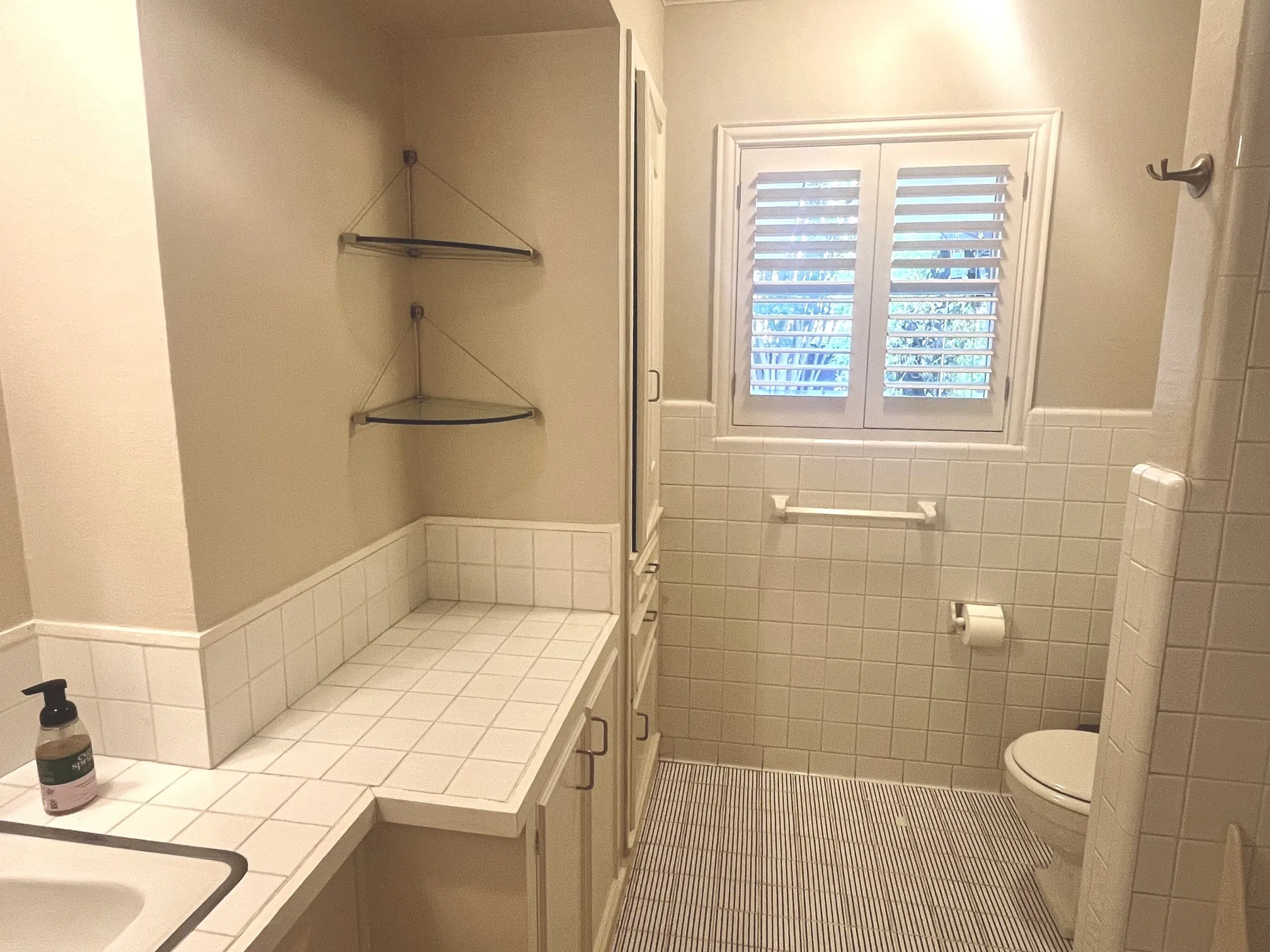 White tile wainscoting and countertop, built-in cabinetry, window with plantation shutters, and glass corner shelving