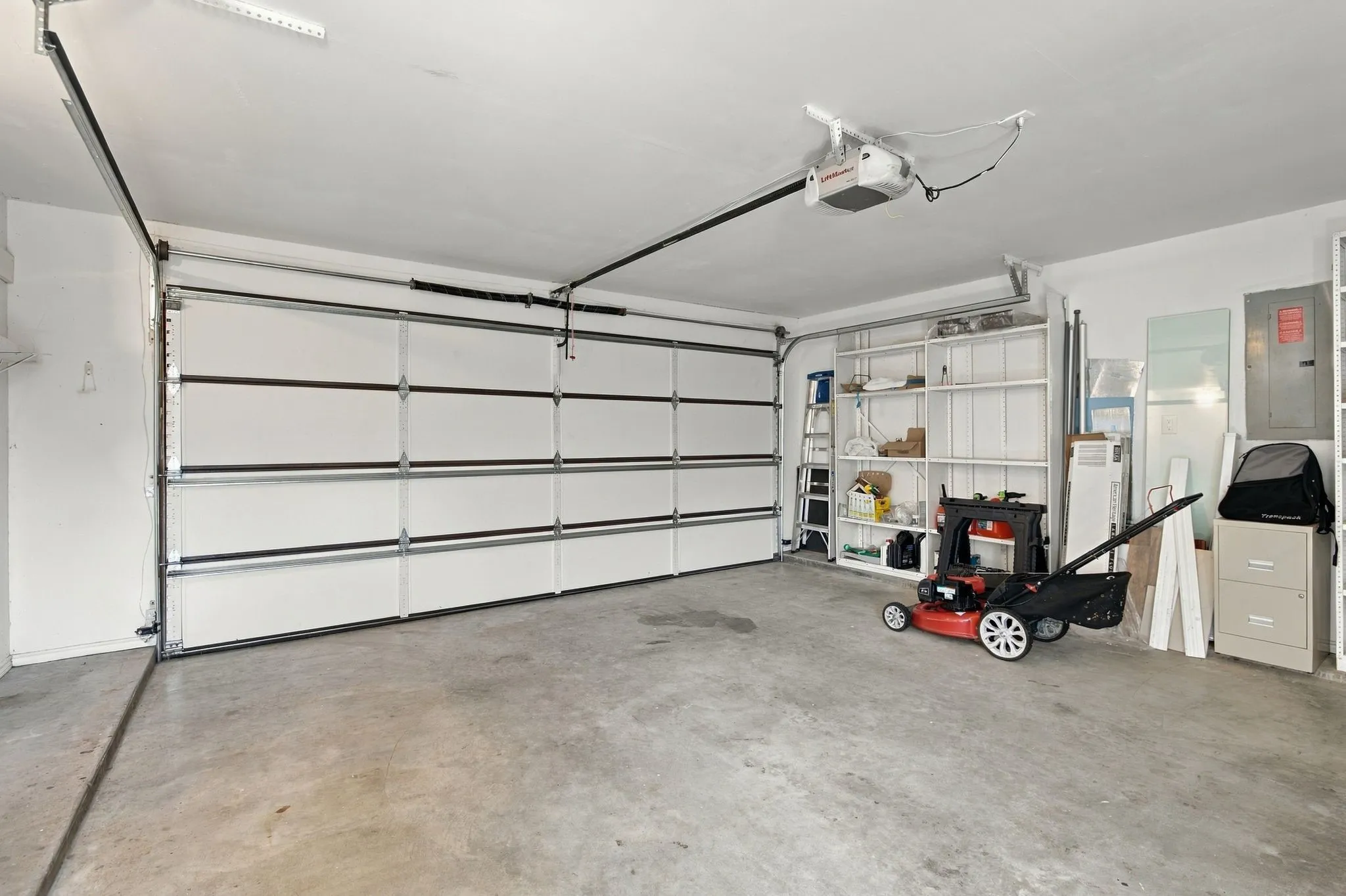 Concrete flooring, white paneled garage door, exposed garage door opener, white painted walls, and built-in shelving