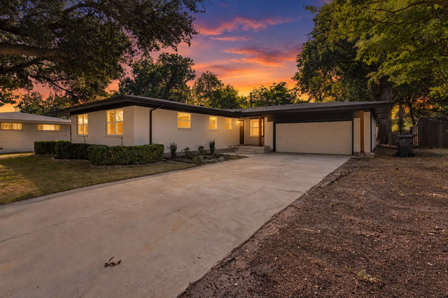 White exterior with dark trim, attached garage with white door, concrete driveway, and mature landscaping