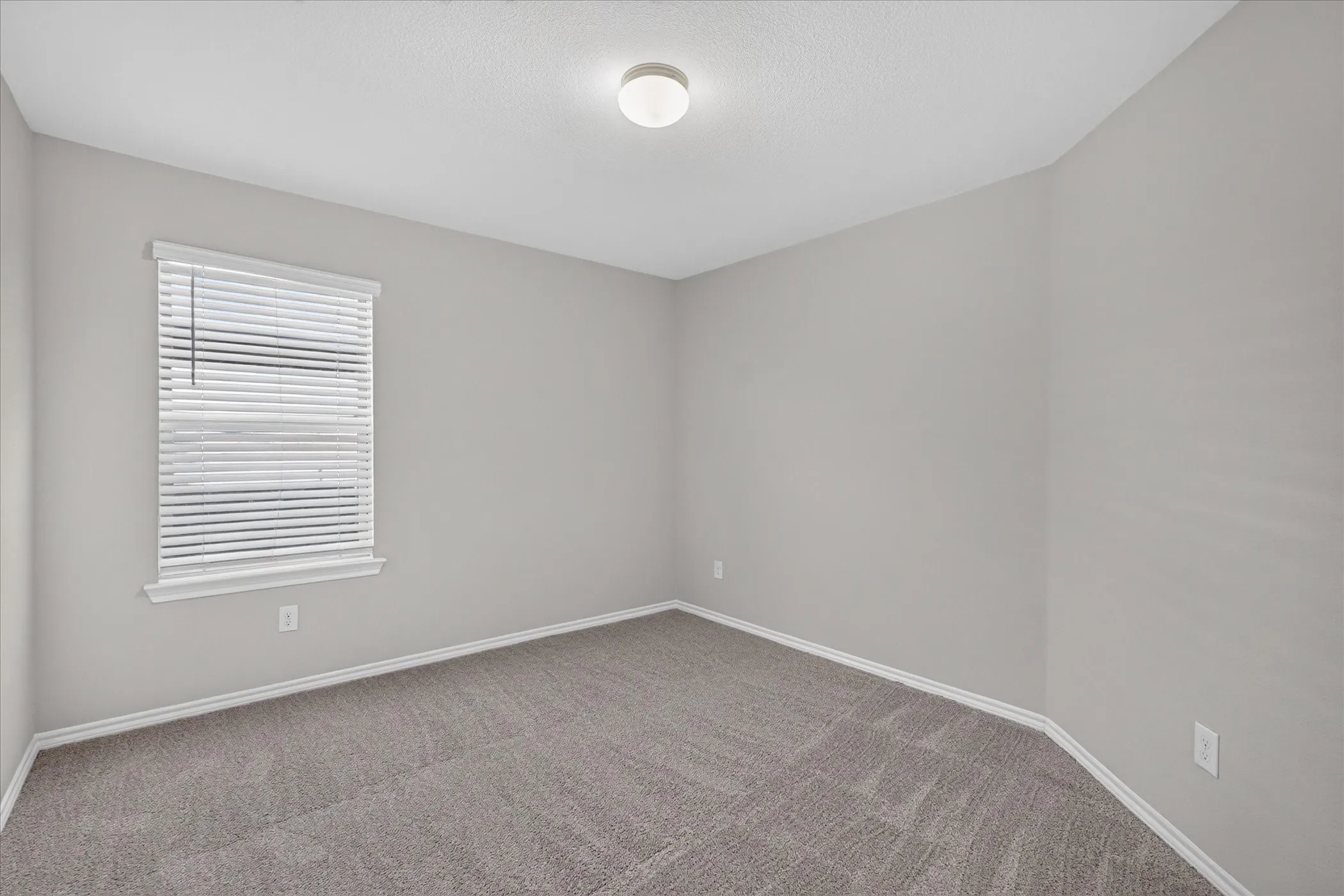 Bright secondary bedroom featuring neutral wall finishes, new carpet flooring with linear pattern detail, large window with horizontal blinds providing natural light, and recessed ceiling lighting