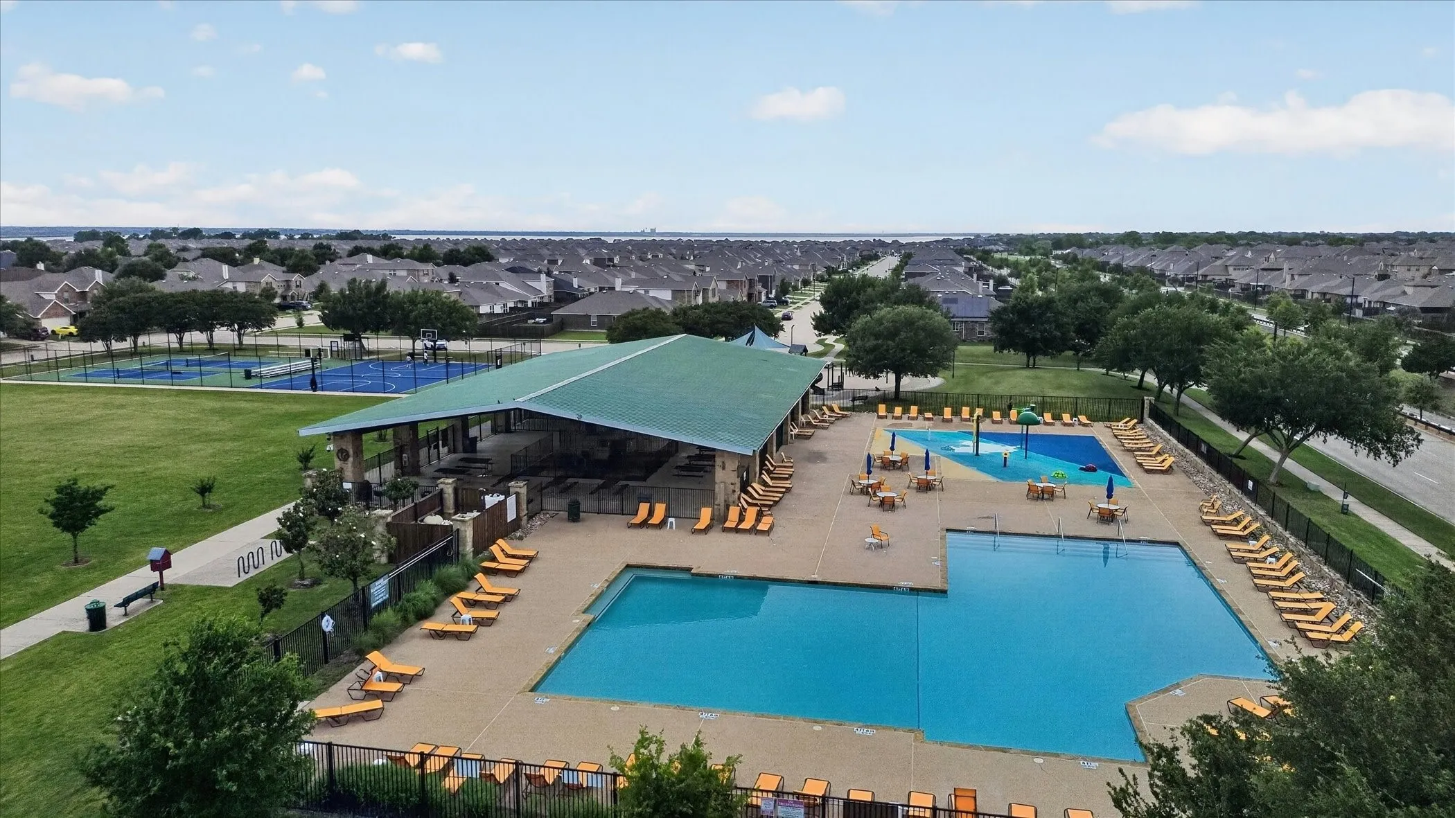 Swimming pool with integrated splash pad, covered pavilion, hardscaped pool deck, adjacent sports courts, and green space