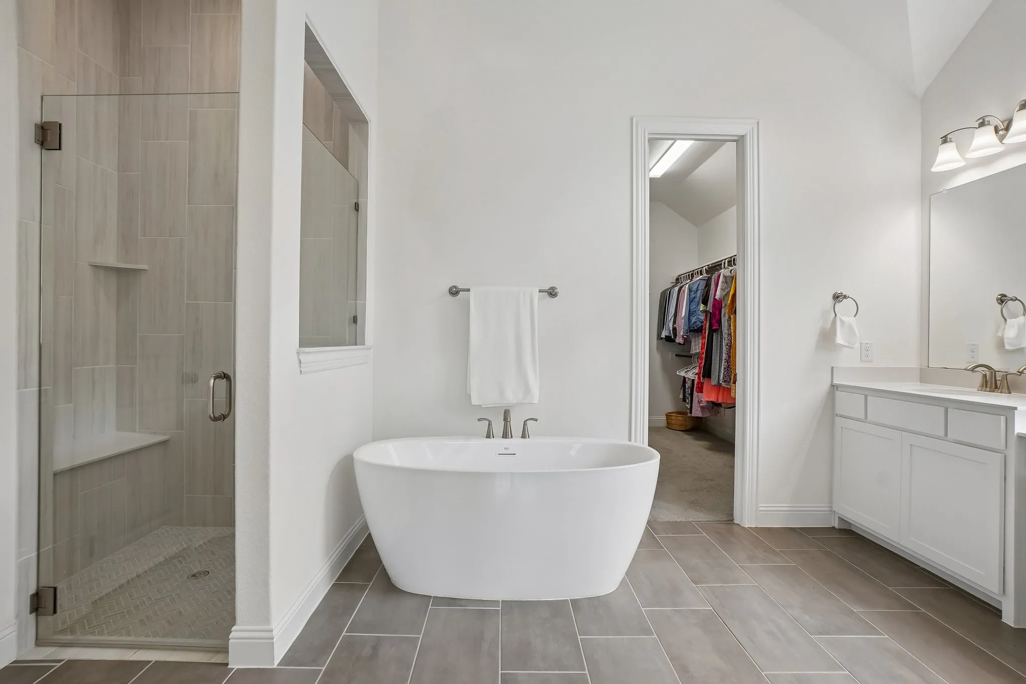 Glass-enclosed shower with a built-in bench, freestanding soaking tub, dual-sink vanity with white cabinetry, and large format floor tile